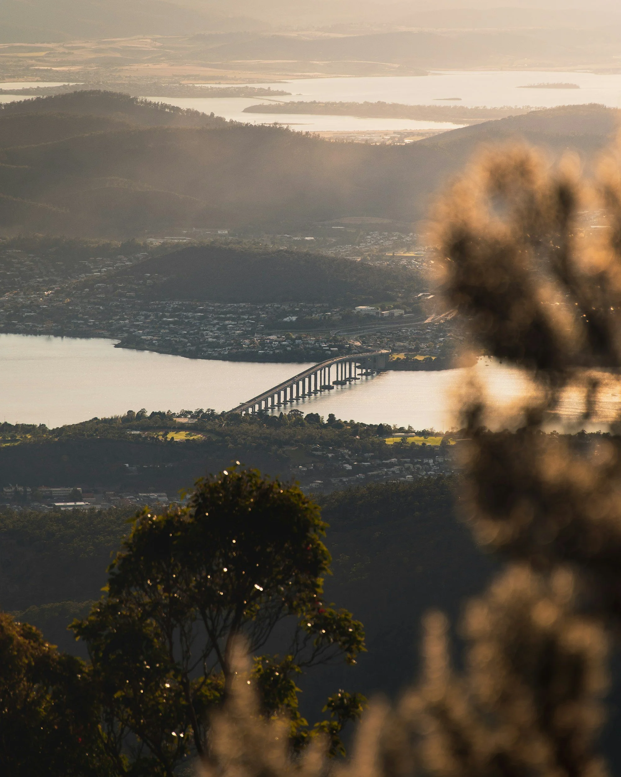 Aerial view of a bridge over a body of water surrounded by hills, with trees in the foreground and mountains in the distance during sunset.