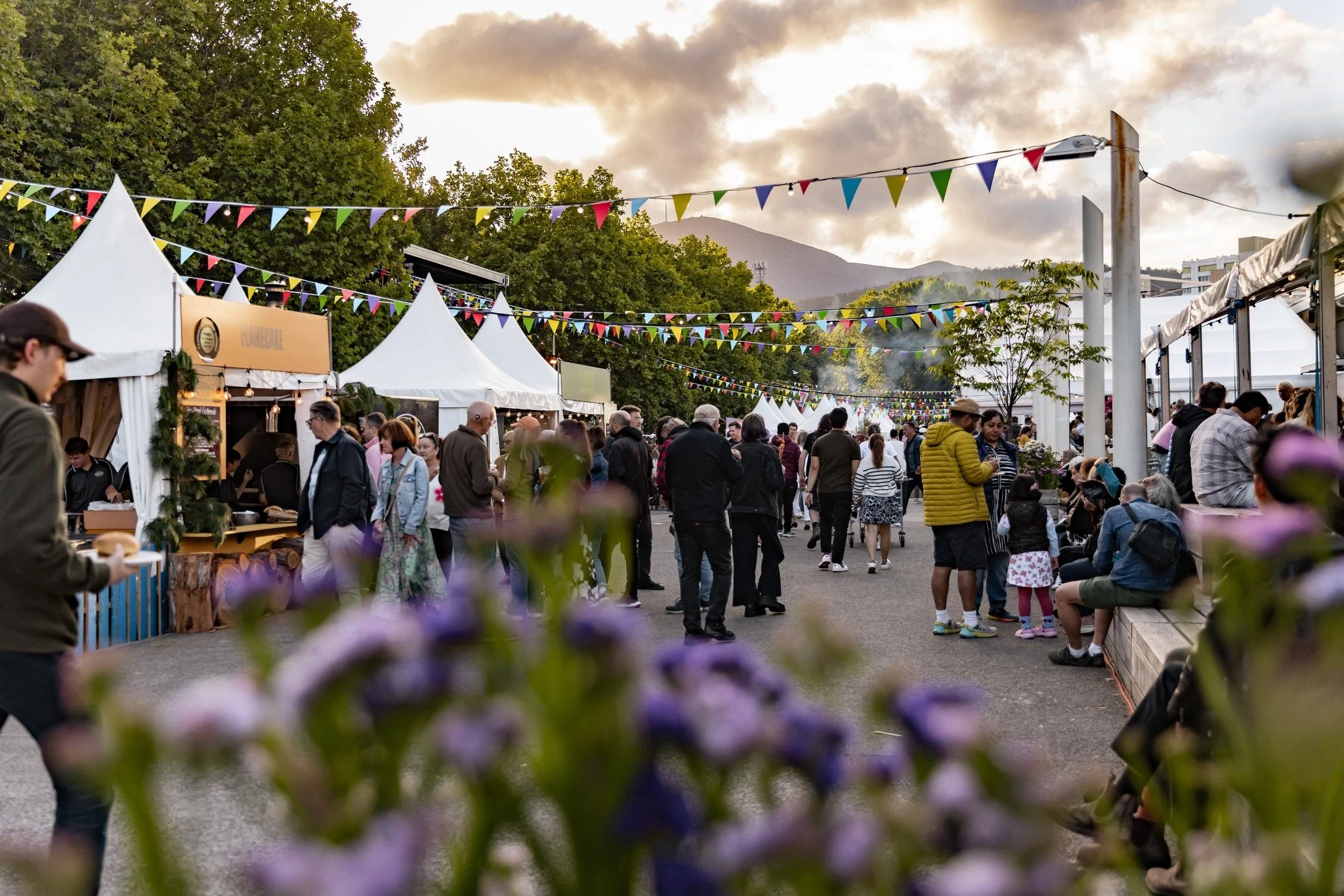 People walking and sitting at an outdoor market with white tents, colorful bunting flags, and mountains in the background during sunset.