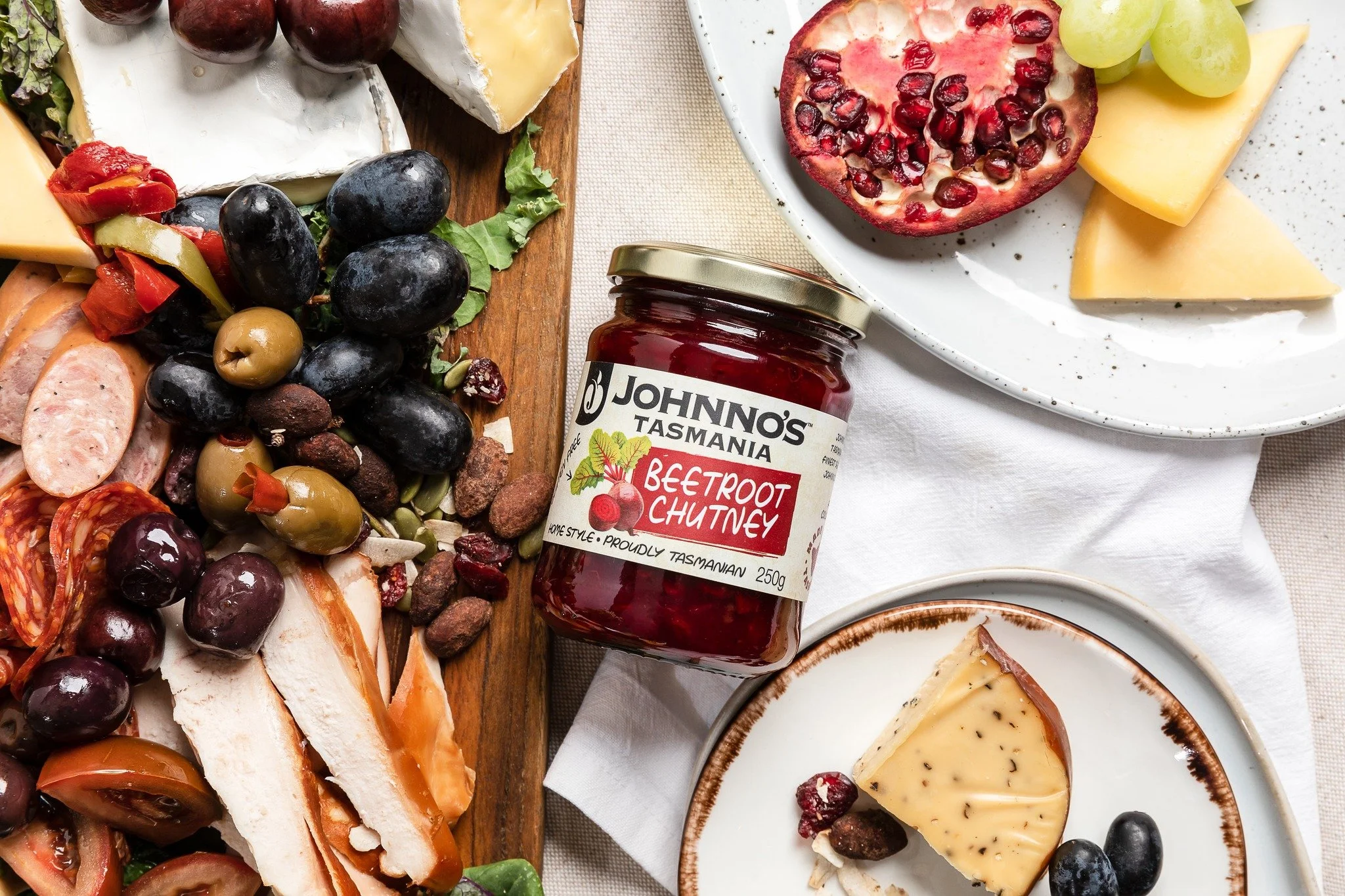 A cheese and meat platter with grapes, olives, and cherry tomatoes, a jar of beetroot chutney, and slices of cheese and pomegranate on plates.