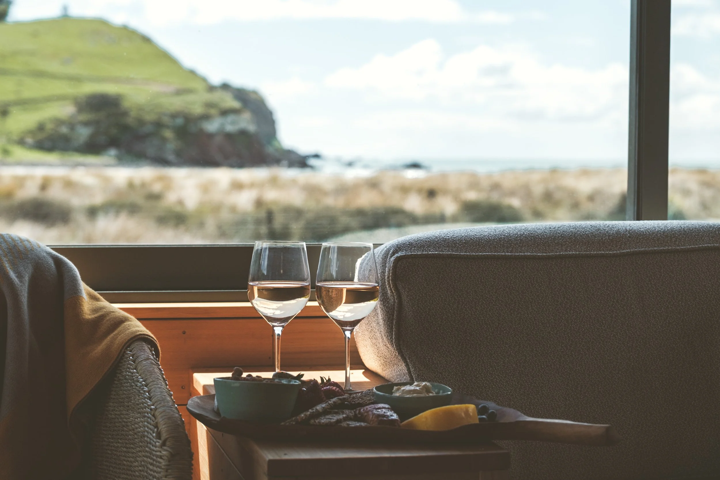 A cozy indoor scene with two glasses of rosé wine, a tray of snacks including cheese, fruit, and nuts, near a large window showing a rocky coast and hillside in the background.