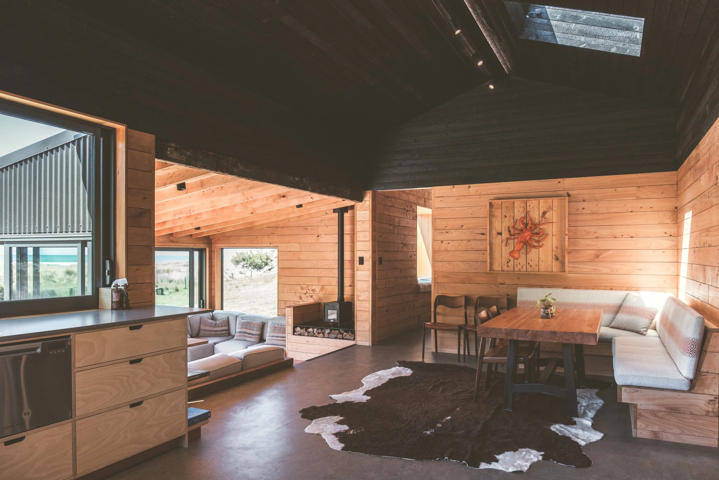 Interior of a cozy cabin with wooden walls and ceiling, featuring a window overlooking greenery, a wood stove, a dining area with a corner bench and wooden table, a cowhide rug, and a wall art piece of a lobster.