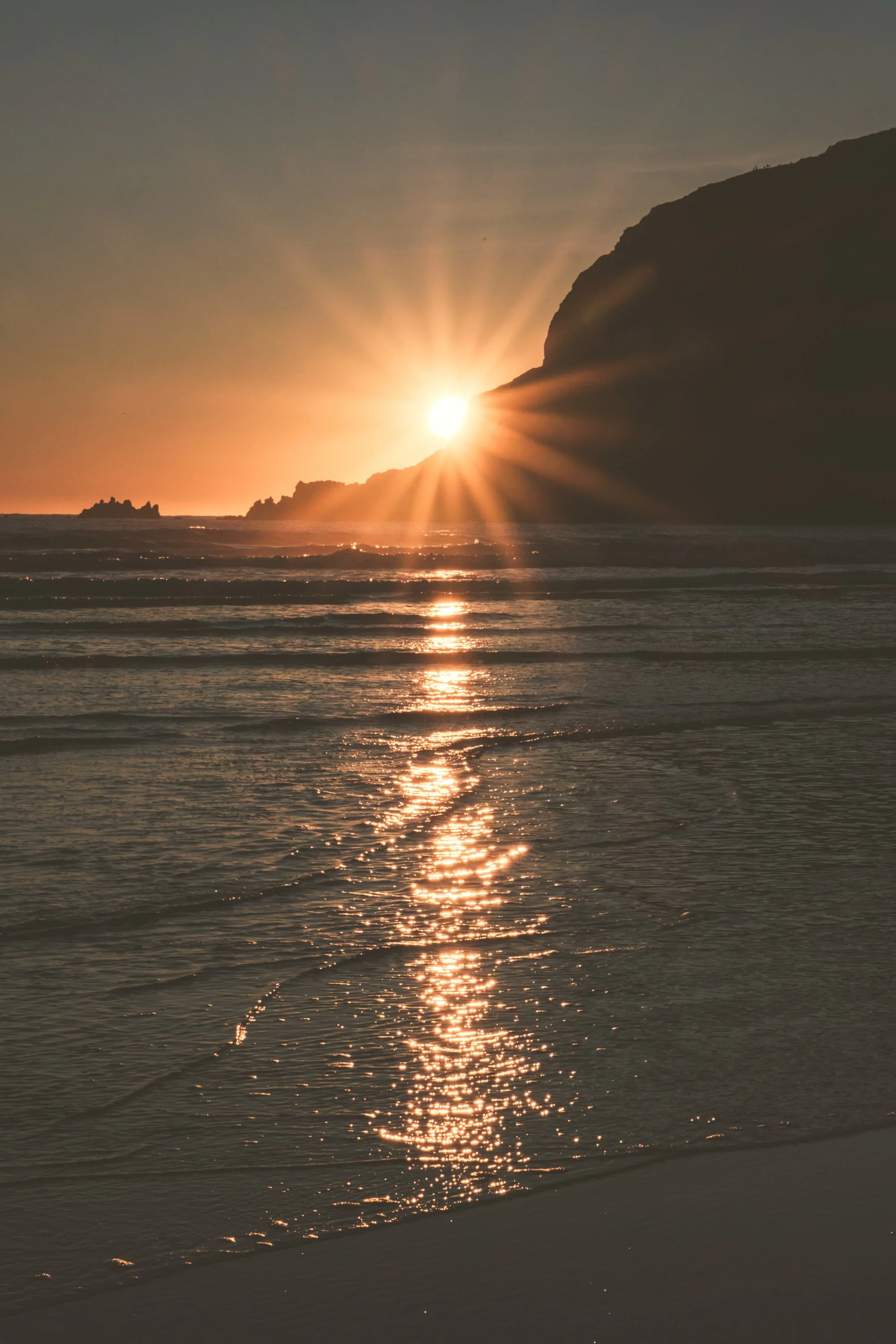 Sunset over the ocean with light reflecting off the water and a rocky cliff on the right.