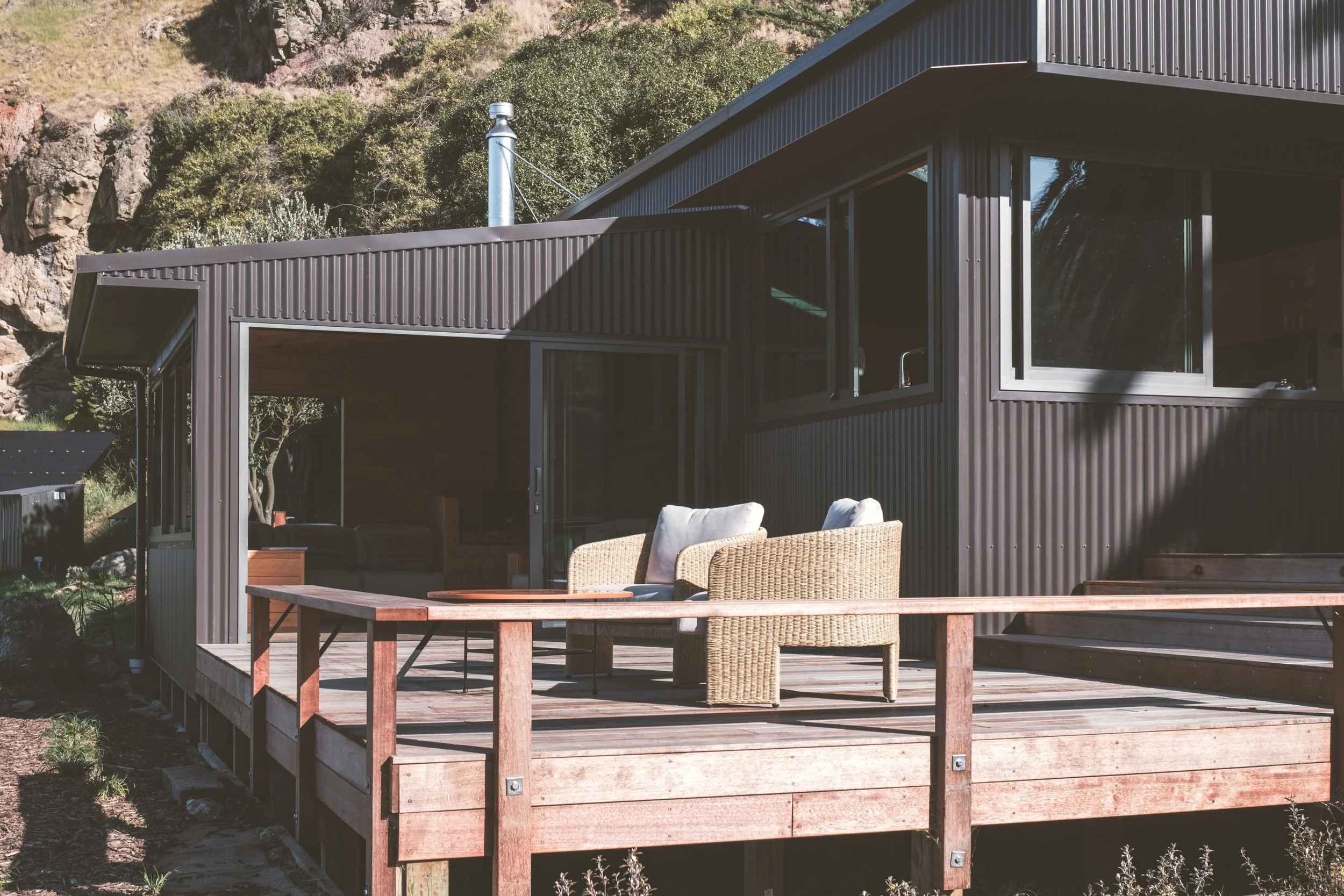 A modern house with black metal siding and large windows, featuring a wooden deck with outdoor seating and a view of surrounding hills and trees.