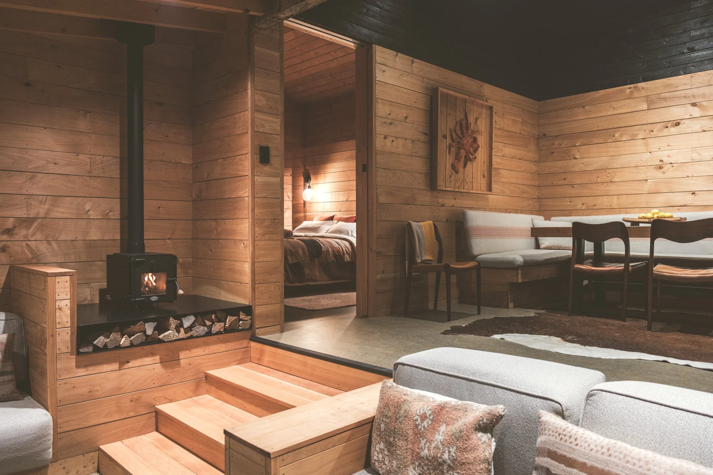 Interior of a cozy cabin living area with wood-paneled walls, a wood stove with stacked logs, a dining table with chairs, and a seating area with a sofa and cushions, leading to a bedroom in the background.