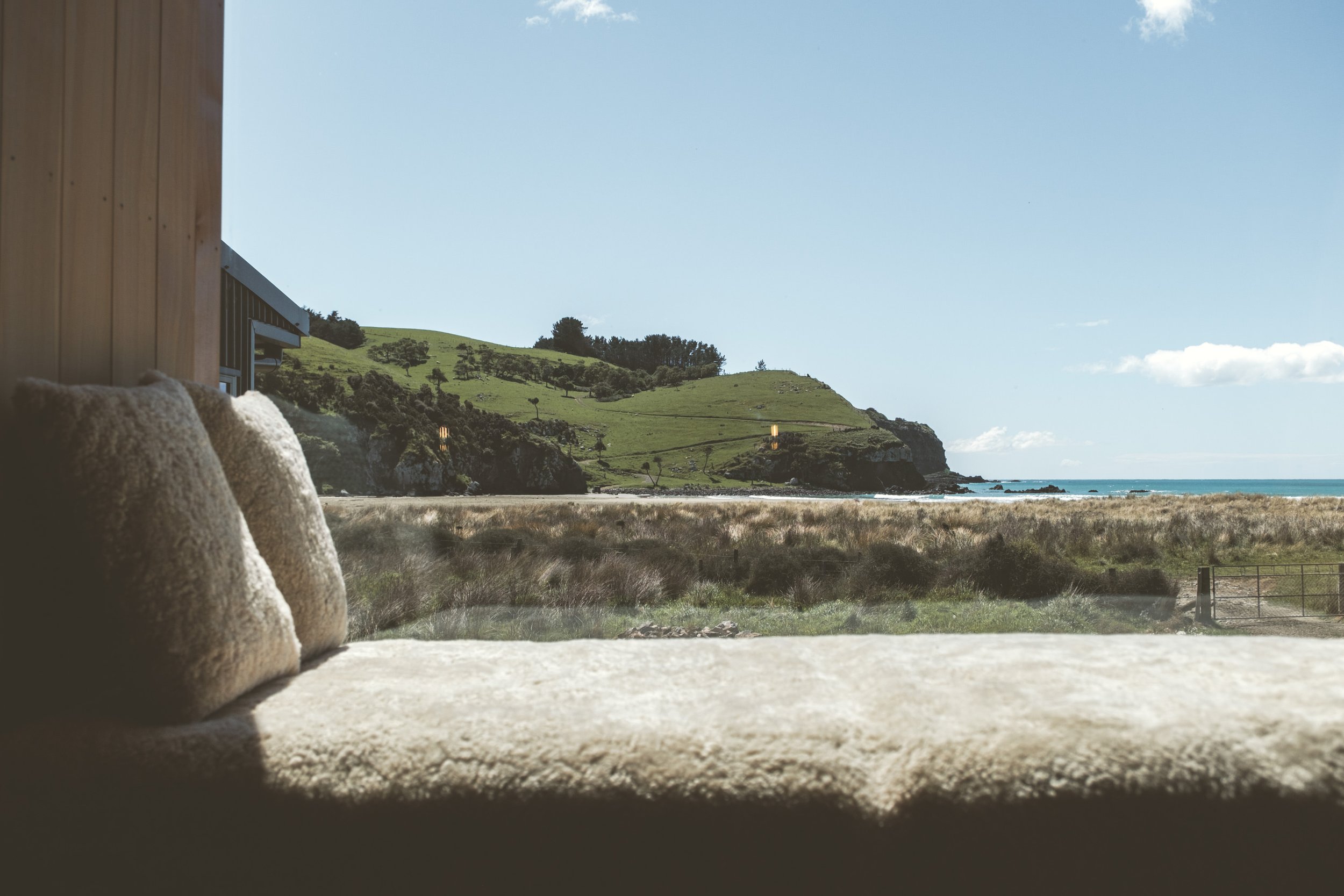 View through a window showing a grassy hill, rocky coastline, ocean, and sky.