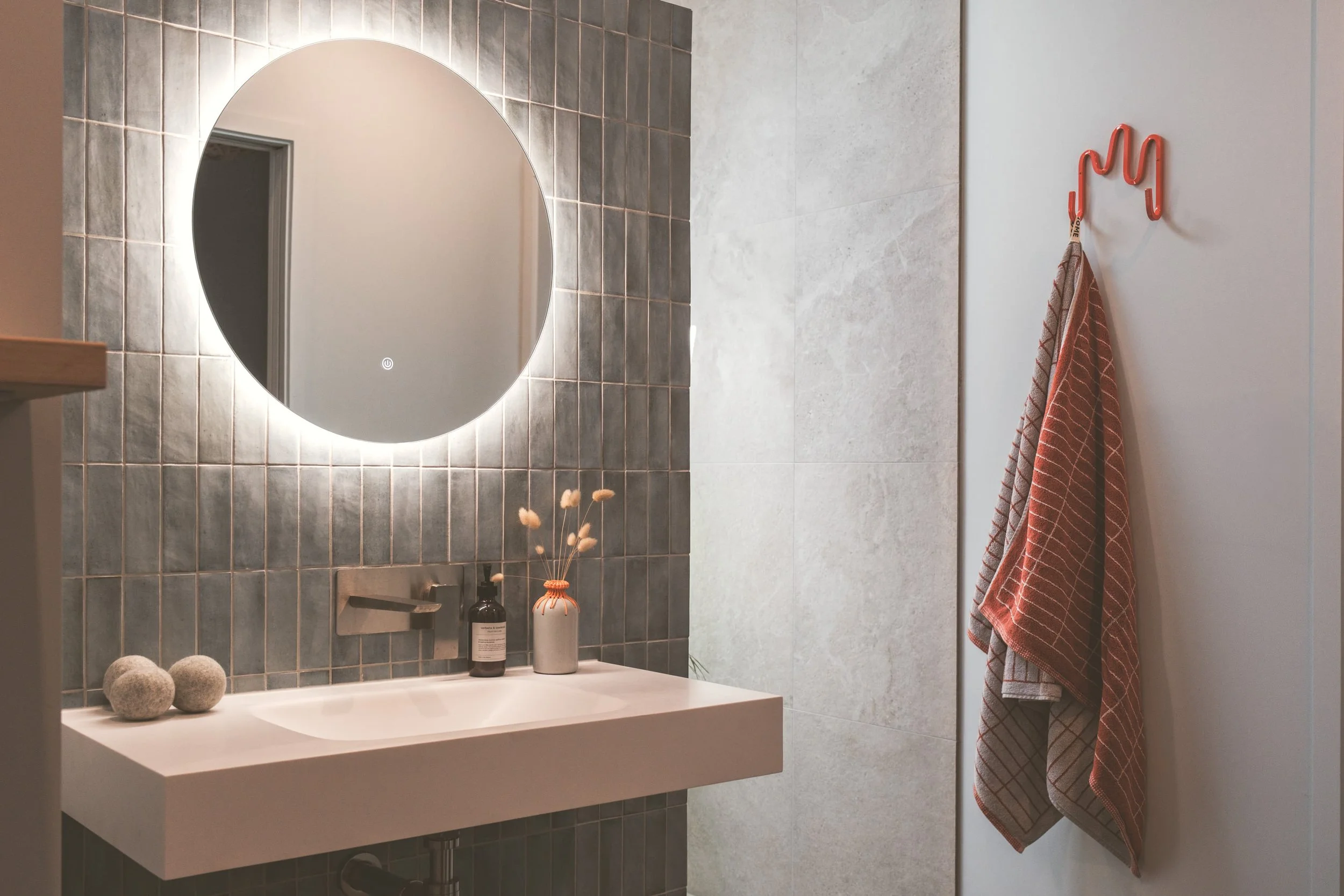Modern bathroom with illuminated round mirror, grey tile wall, white sink, black soap dispenser, decorative vase with dried flowers, and striped towels hanging on a pink hook.