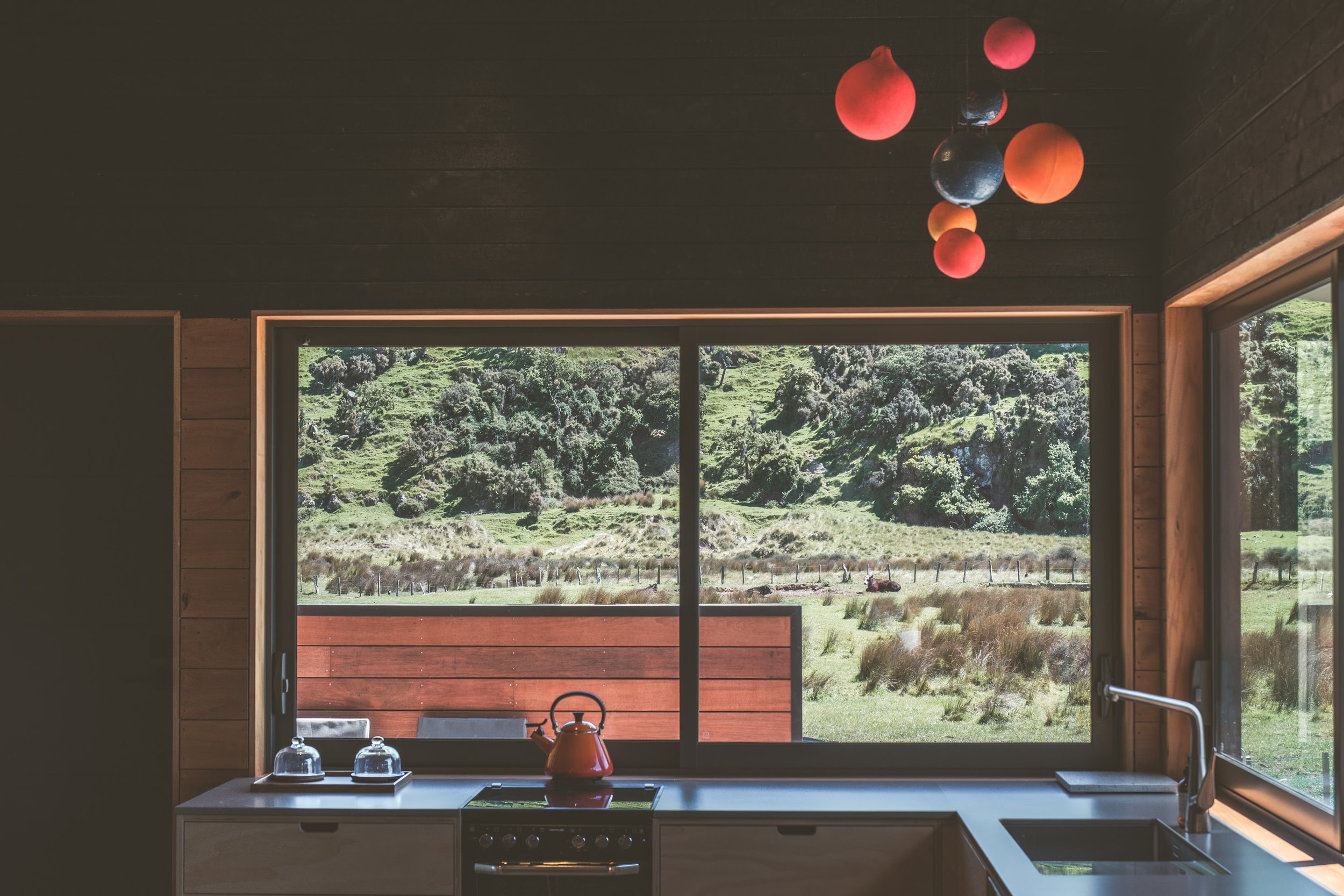 A kitchen with large window overlooking a green landscape with grass, bushes, and trees. The kitchen counter has a red teapot, two glass jars, and a black stove. An above-head colorful light fixture hangs from the wooden ceiling.