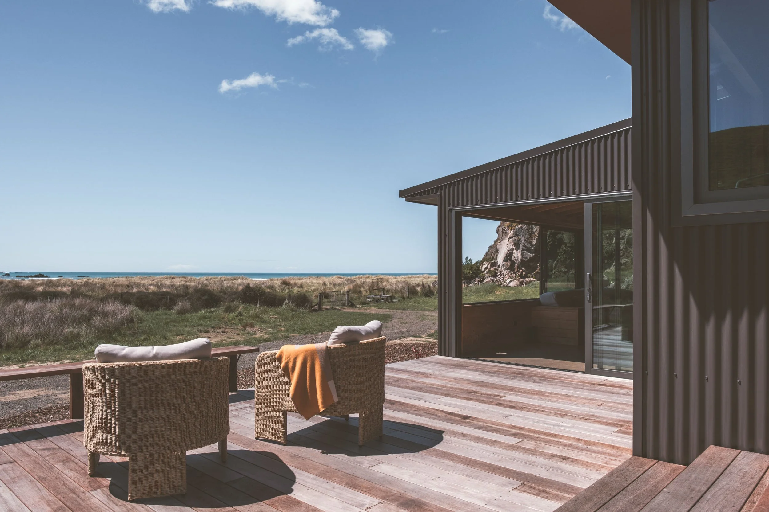 Beachside house with a wooden deck, wicker chairs with cushions and a blanket, and ocean view with grass and rocky landscape