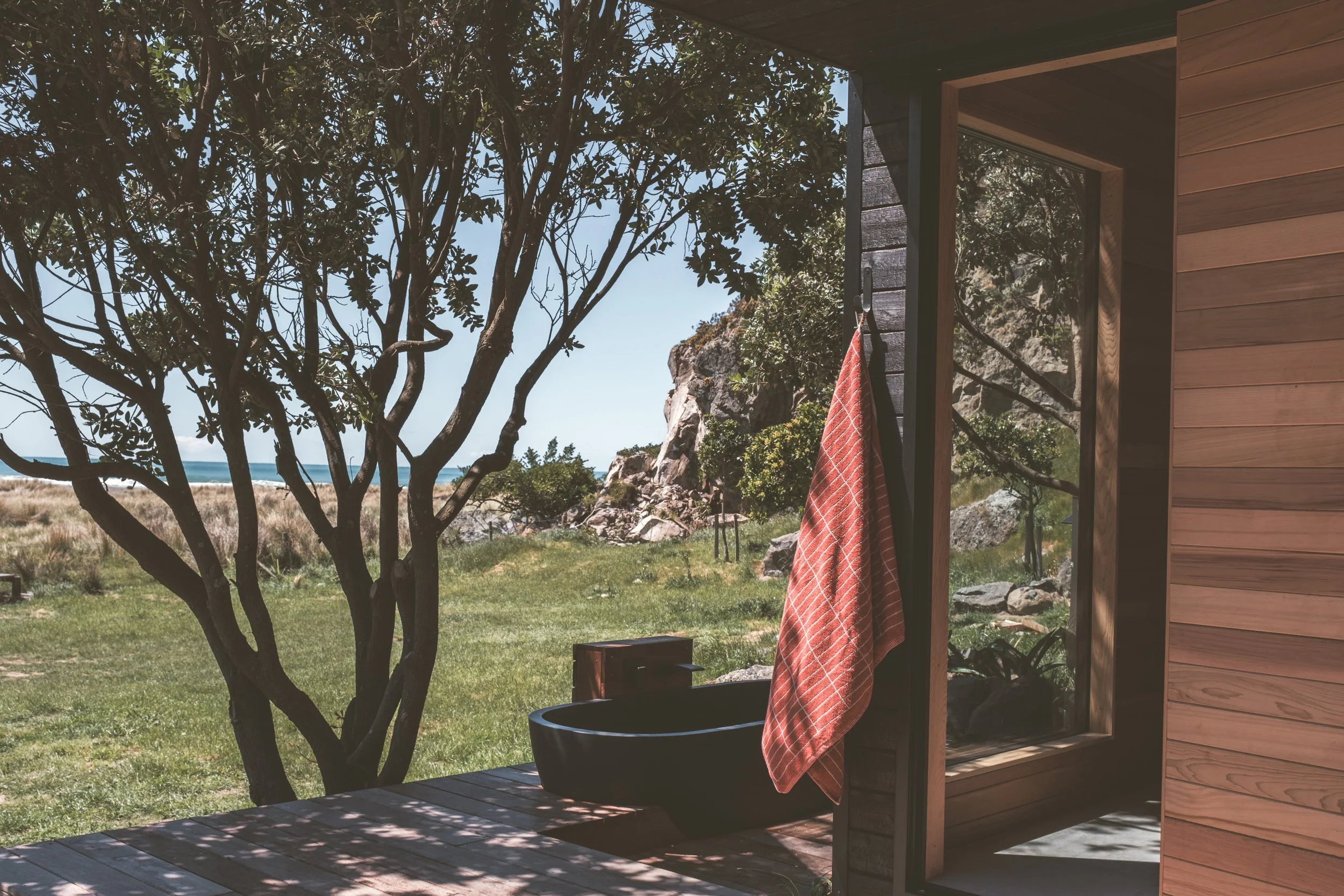 Outdoor scene with a wooden building, a large tree, grass, rocks, and the ocean in the background. There is a black bathtub outside, with a red towel hanging on the wall beside a window.