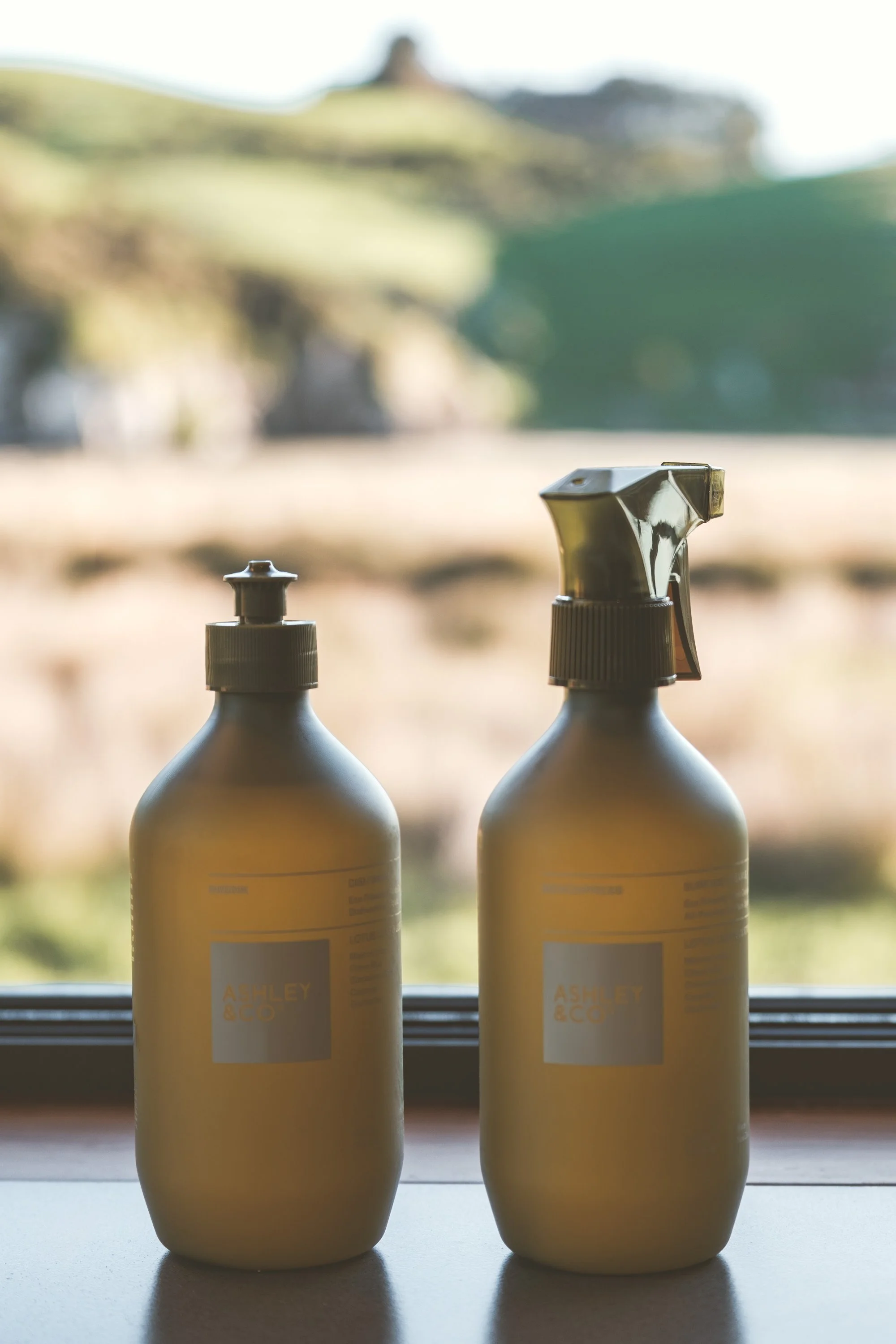 Two beige spray bottles labeled Ashley & Co placed on a windowsill, with a blurred outdoor landscape in the background.