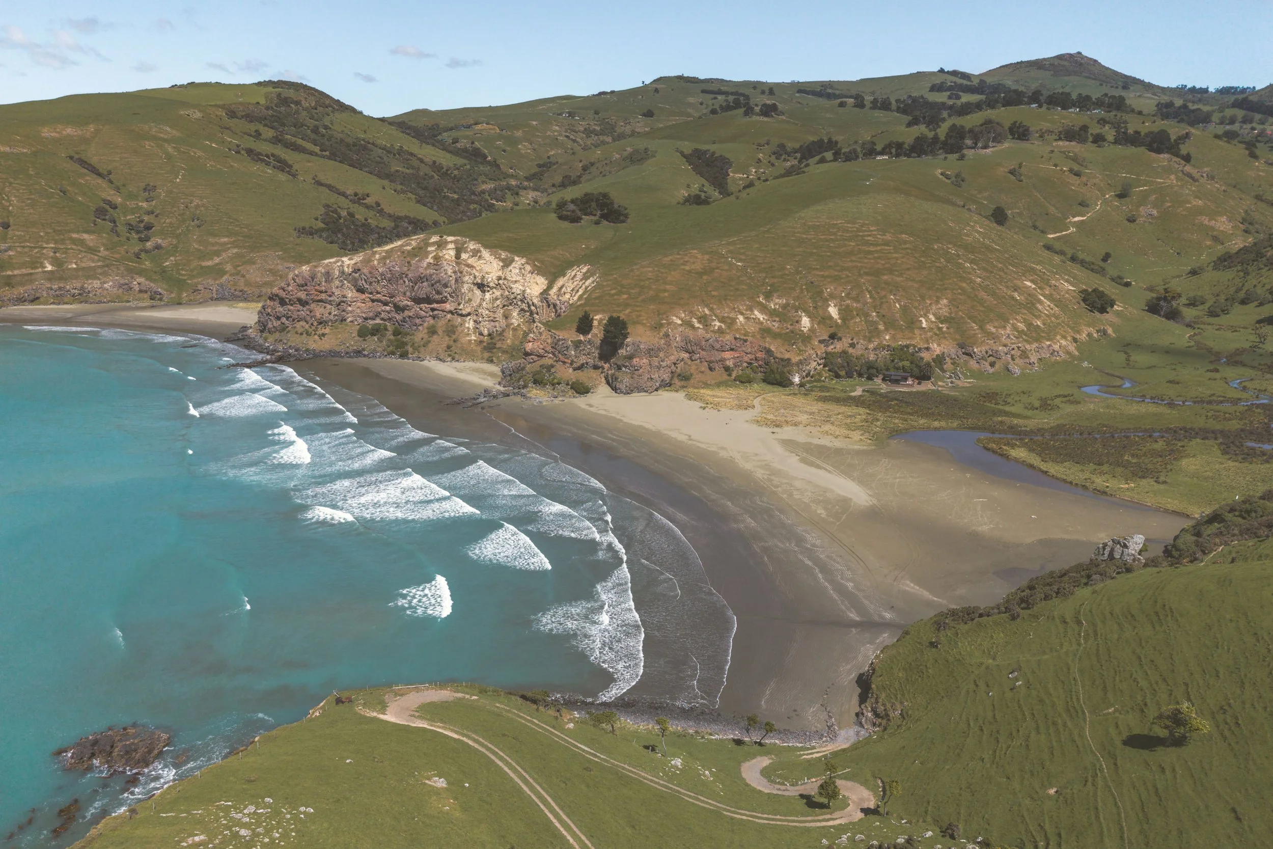 Aerial view of a beach with turquoise water, walking paths, green hills, and cliffs.