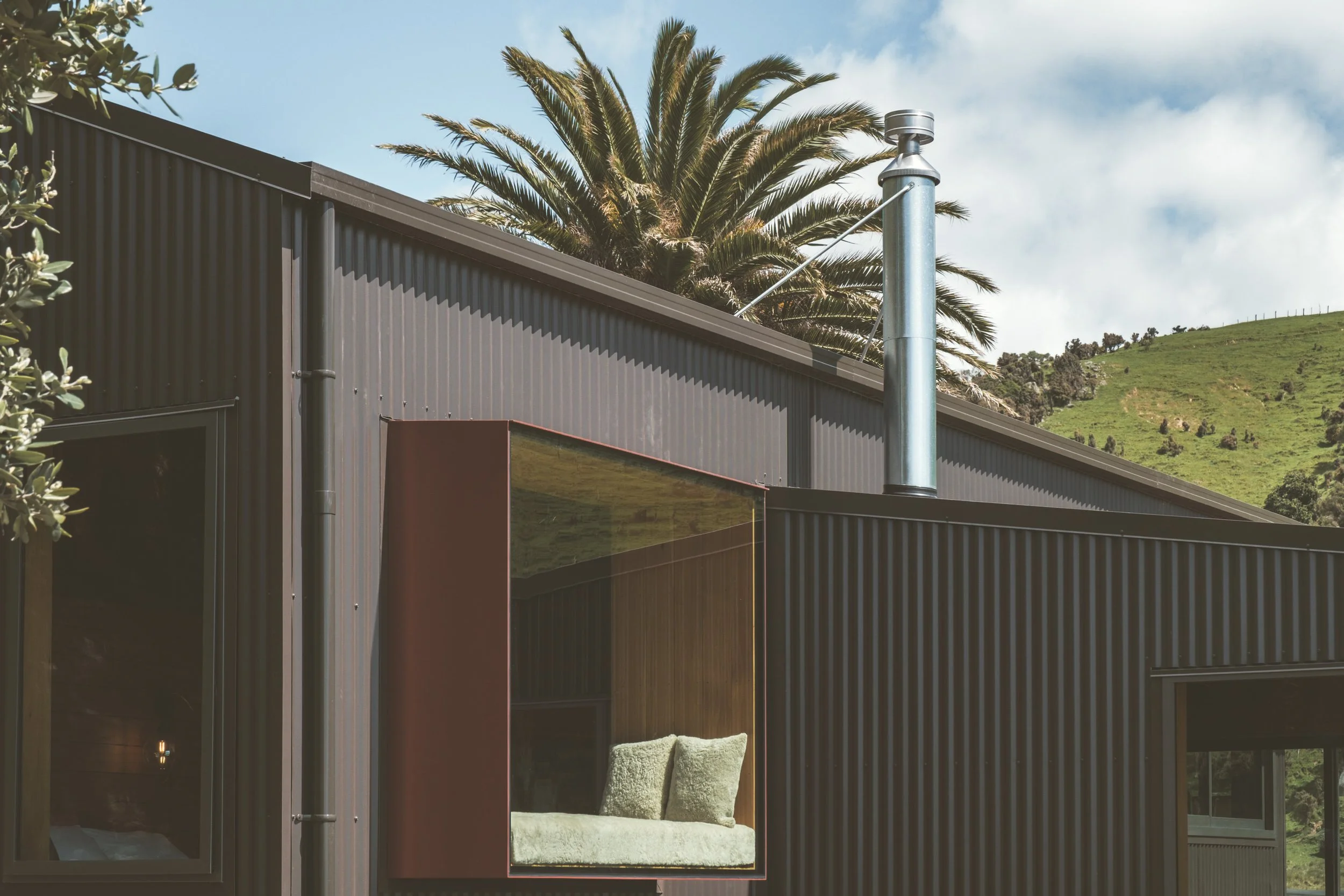 Close-up of a modern house with black corrugated metal siding and a large window with a view of a green hillside and palm trees in the background.