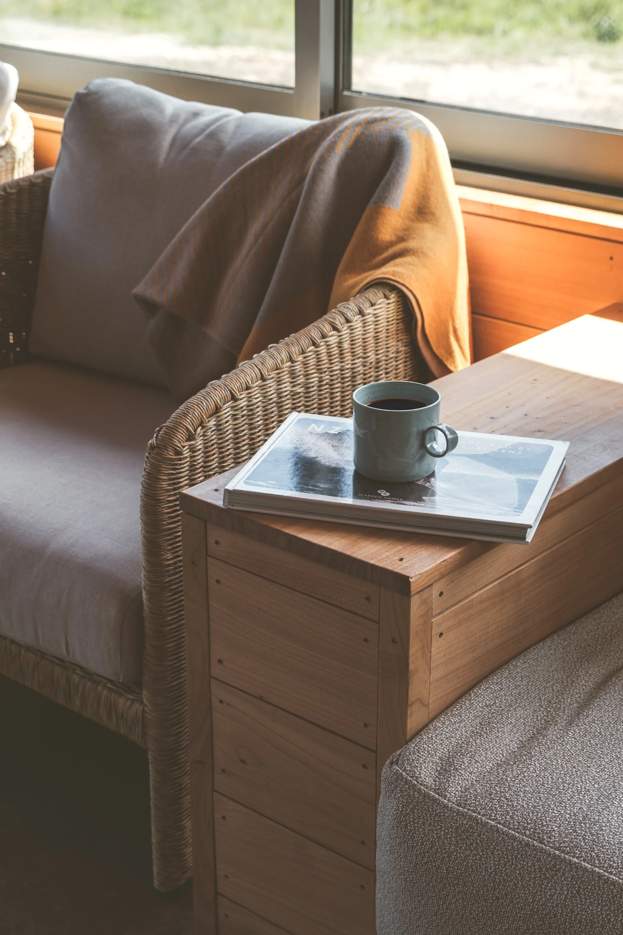 A cozy interior scene with a wicker sofa, a wooden side table, a coffee mug, and a magazine, next to a window letting in natural light.