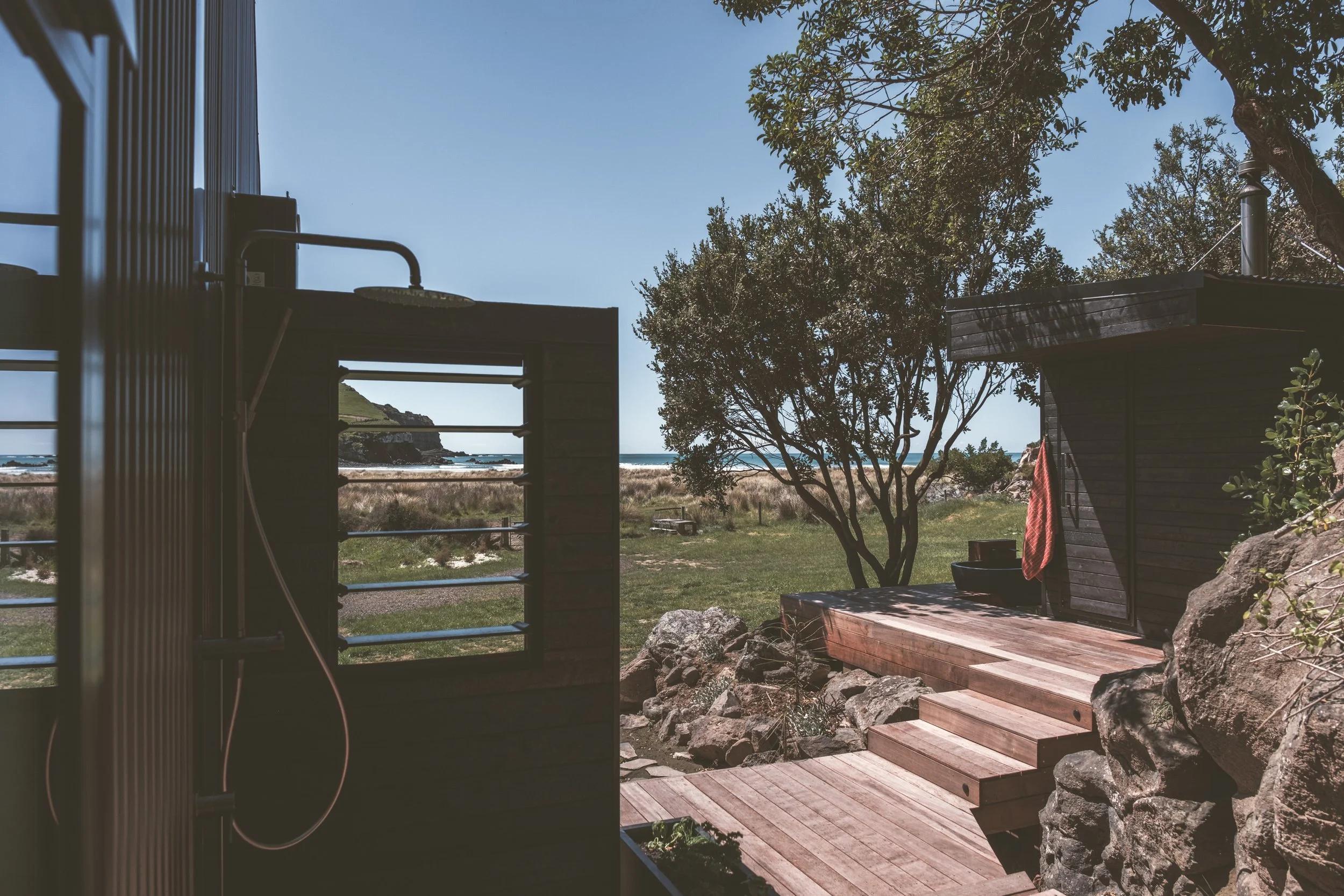 Outdoor shower on a wooden deck with a view of the beach, ocean, and sky, surrounded by trees and rocks.