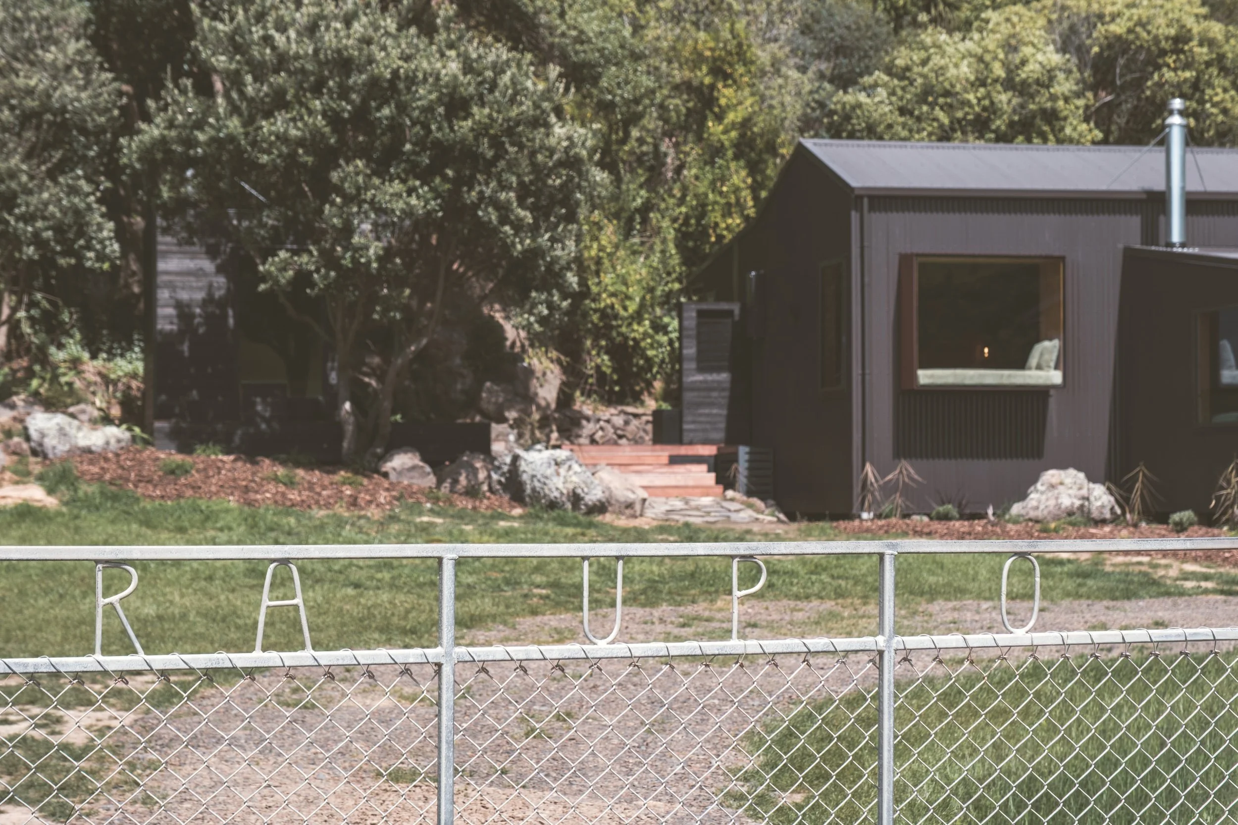 A chain-link fence with metal letters spelling 'RAUP' in front of a grassy area with a modern dark house and trees in the background.