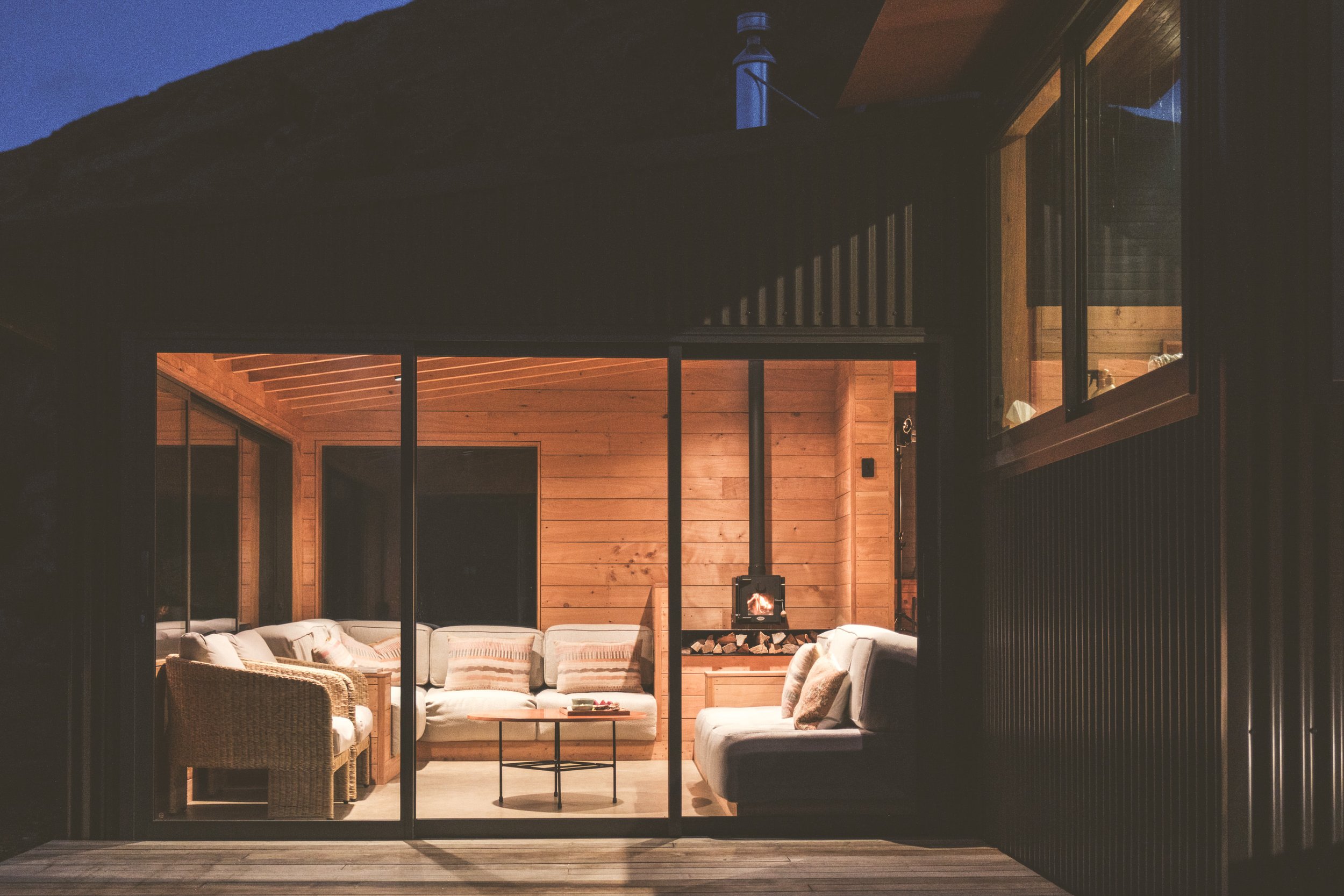 Night view of a cozy living room through glass sliding doors, featuring a wood-paneled interior with sofas, a wicker chair, a small round coffee table, and a fireplace with logs, illuminated warmly inside.