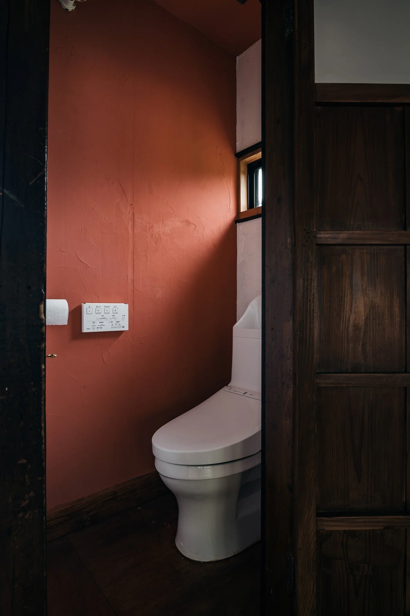 木製の扉の向こうにあるトイレ室内の白い便器と壁に取り付けられた操作パネルとペーパーホルダーが見える