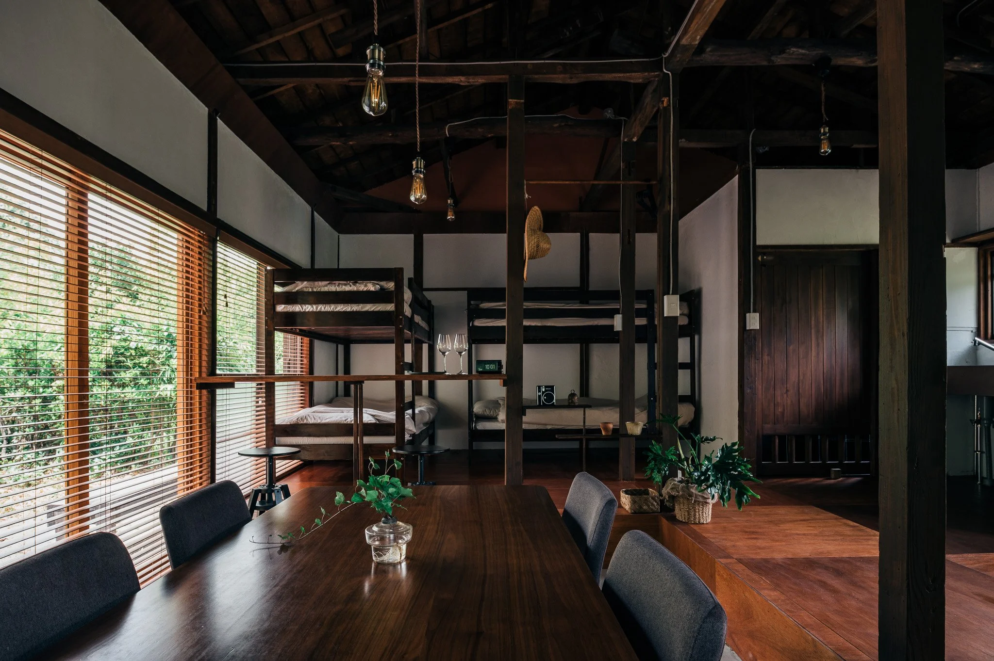 木製のシンプルな家具と観葉植物が飾られた居心地の良いリビングスペース。大きな窓から自然光が差し込み、木のブラインドが窓を覆っている。上階にロフトベッドと小さなテーブルが見える。