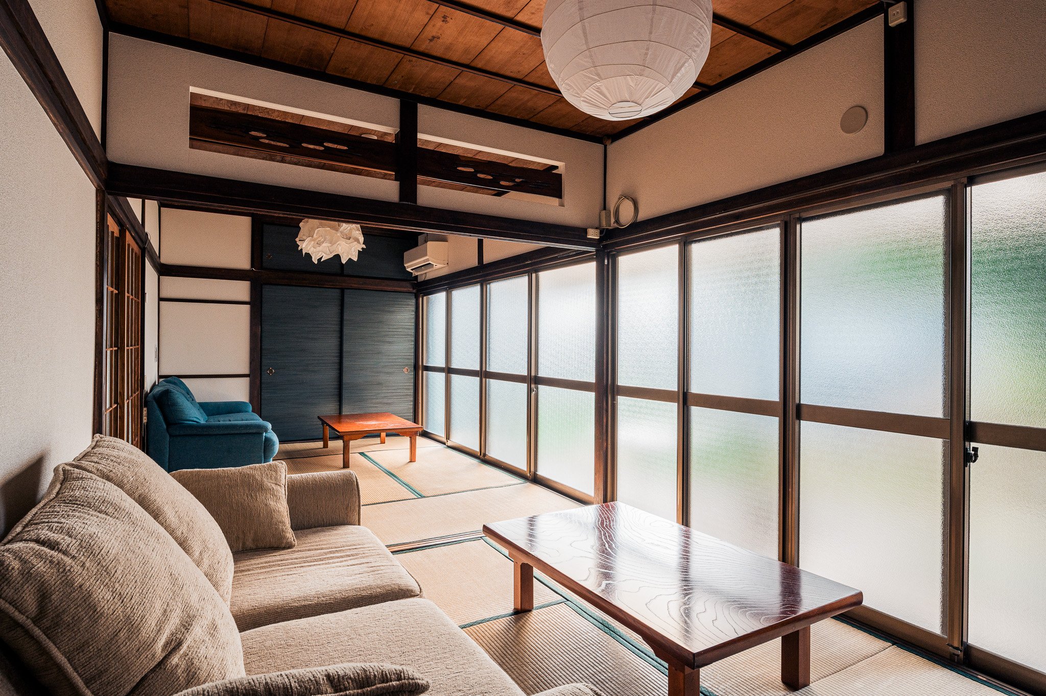 伝統的な和風のリビングルームで、ソファ、低い木製のテーブル、座布団が敷かれた畳の床、大きな障子窓と紙製のランプシェードがある。