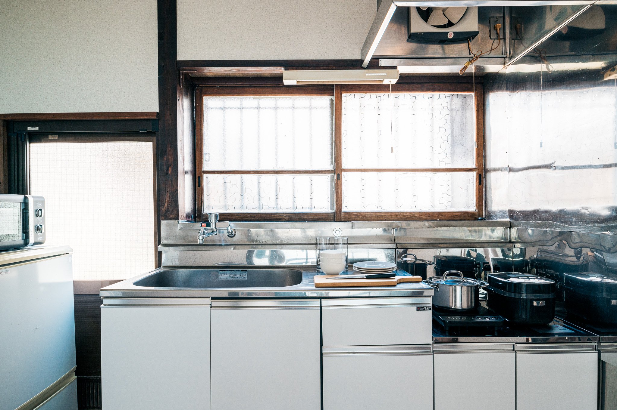 シルバーのステンレス製キッチンシンクとガスコンロ、窓と電子レンジや冷蔵庫が映るキッチンの内部写真