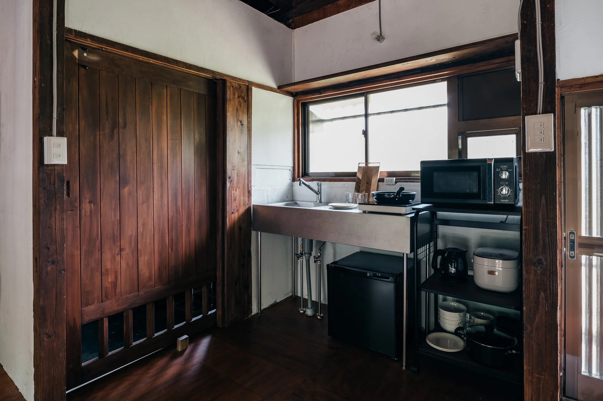 木製の扉と窓がある小さなキッチン。シンク、電子レンジ、炊飯器、電気ケトル、食器類が置かれている。
