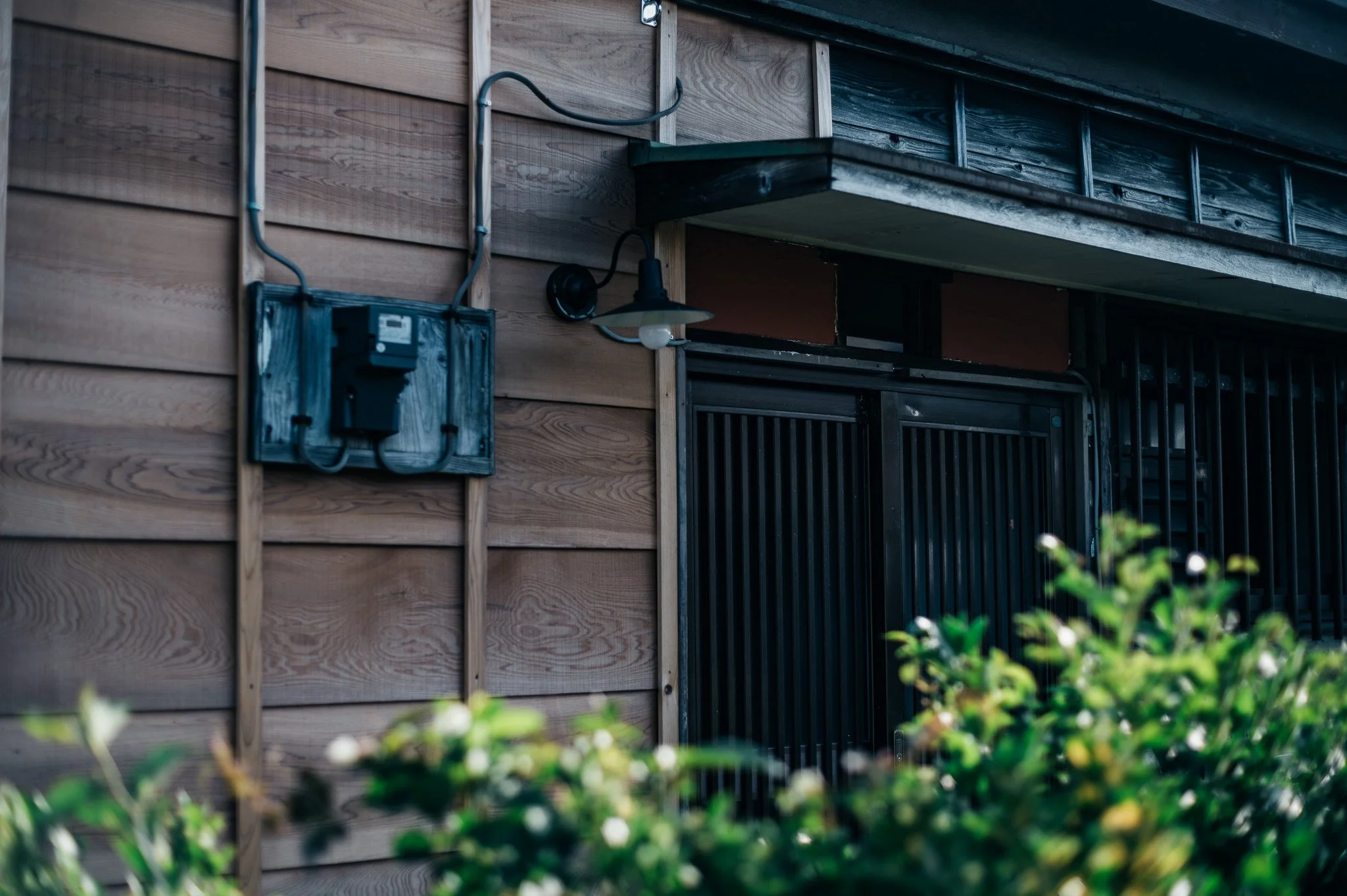 木製の建物の壁面に電気の配線と黒い電気メーター、照明器具が設置されている。前景には緑の植物が見える。