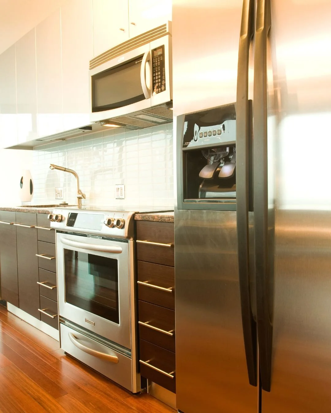 Modern kitchen with stainless steel refrigerator, oven, microwave, and coffee maker. Dark wood cabinets and white tile backsplash with a sink and faucet.