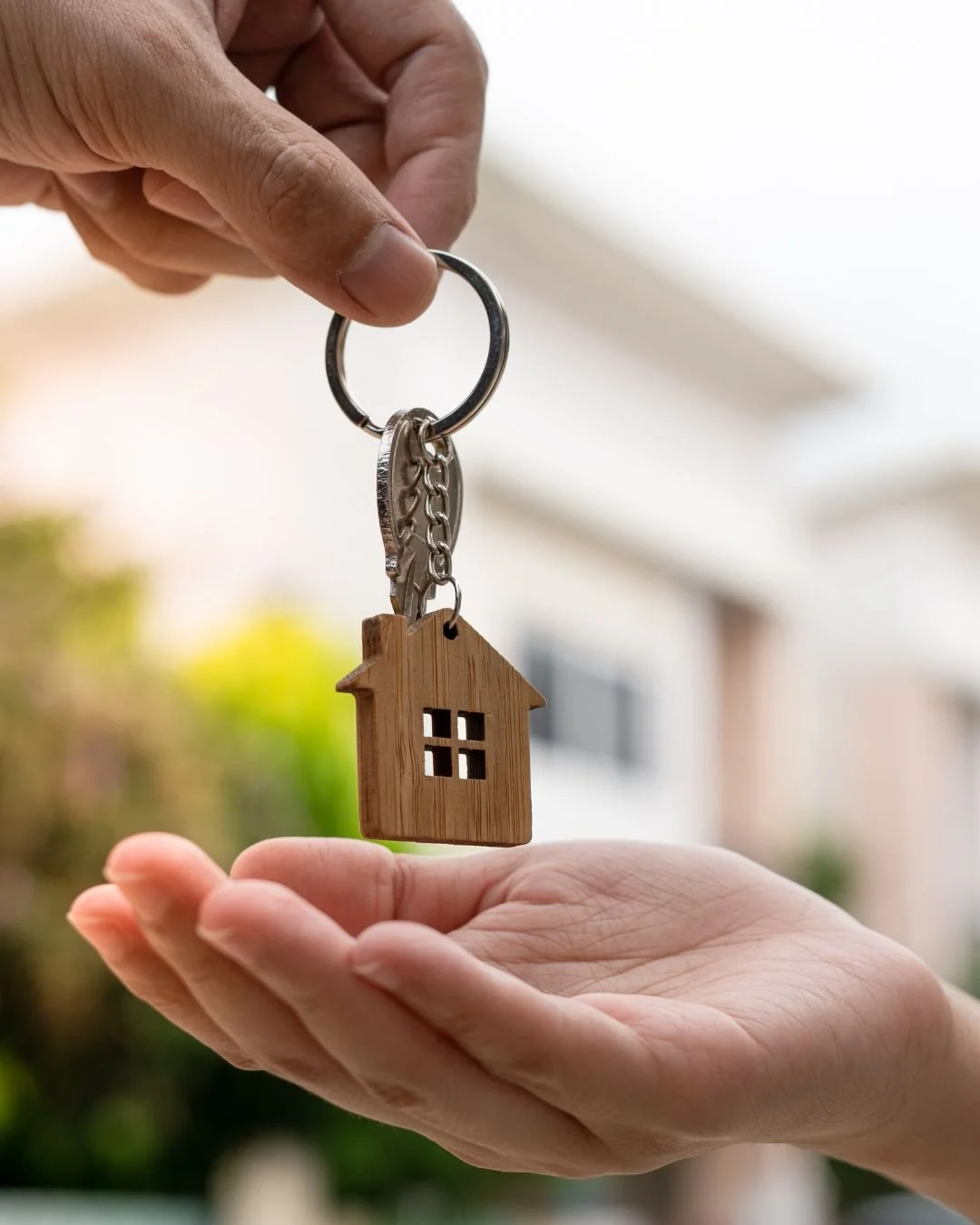 A hand is holding a keyring with a small wooden house-shaped keychain, extended toward another hand.