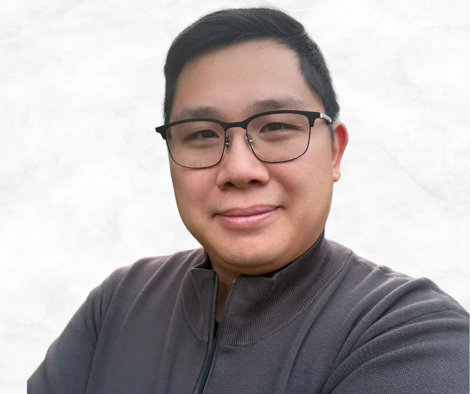 A young man with short black hair, wearing glasses and a dark zip-up jacket, smiling at the camera against a plain white background.