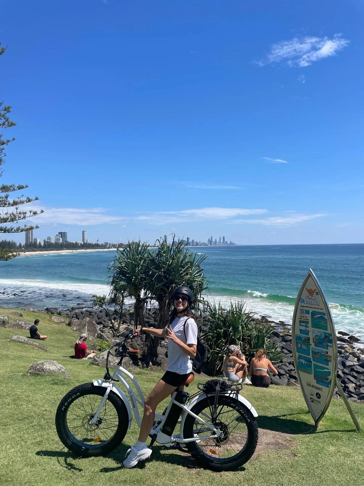 A woman standing beside an electric bike near a coastal park, with people relaxing on the grass and rocks, and a city skyline in the distance on a sunny day.