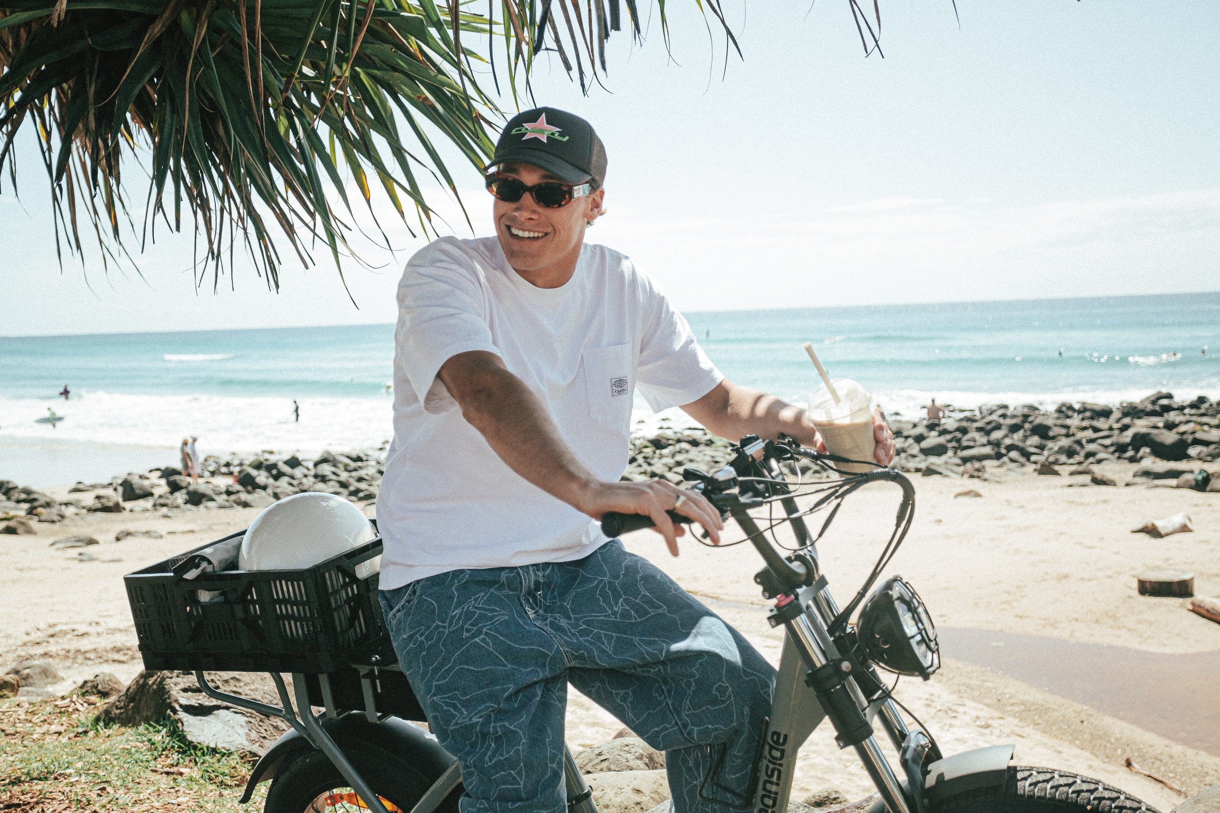 A man riding an electric bike along the beach with a drink in his hand, smiling under a tree with the ocean and rocks in the background.
