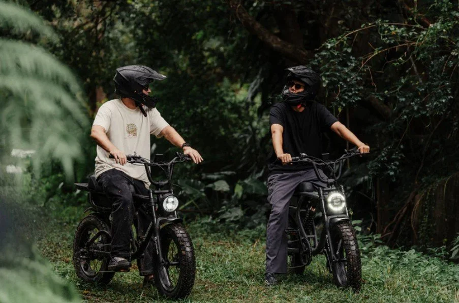 Two individuals riding e-bikes through a dense green forest, both wearing helmets and face masks.