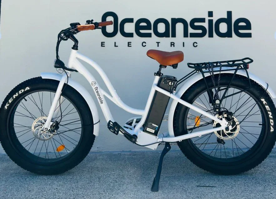 Electric fat tire bicycle with a white frame, brown saddle, and black rear rack, parked outdoors in front of Oceanside Electric signage.
