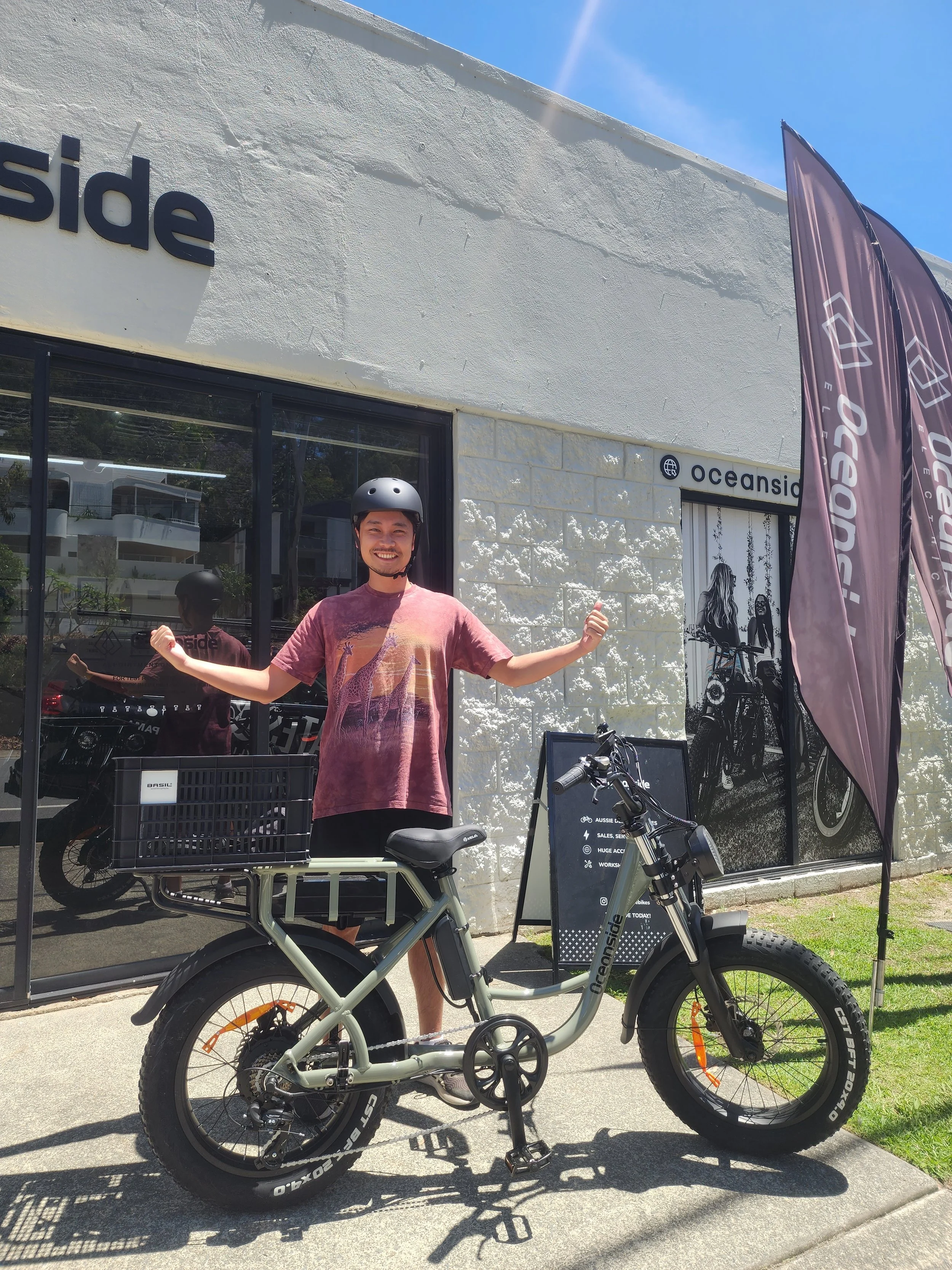 Smiling young man standing next to an electric bike outside a store with flags and signage.