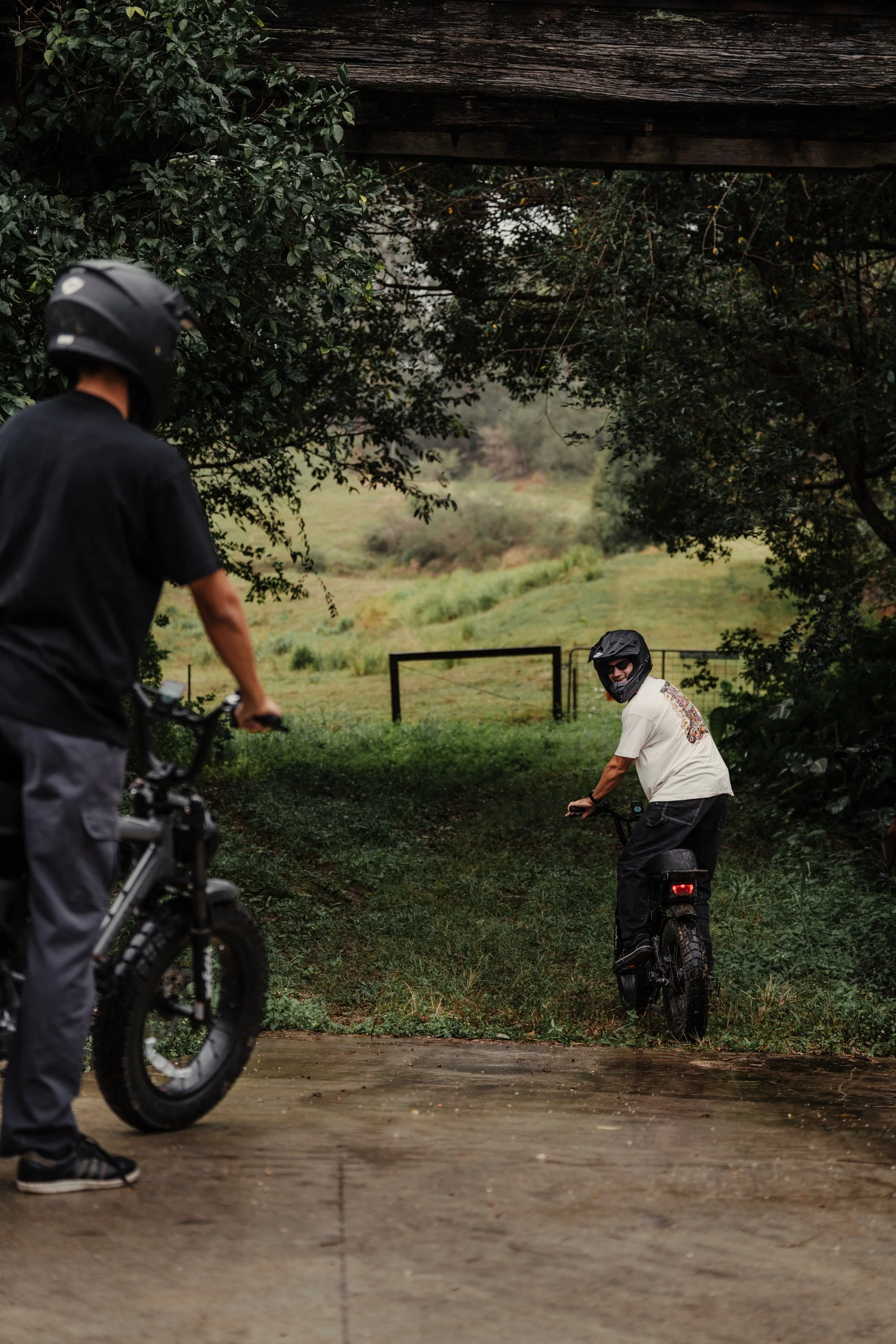 Two men with bikes under a bridge in a green outdoor area during rain. One is facing away, the other is smiling at the camera.