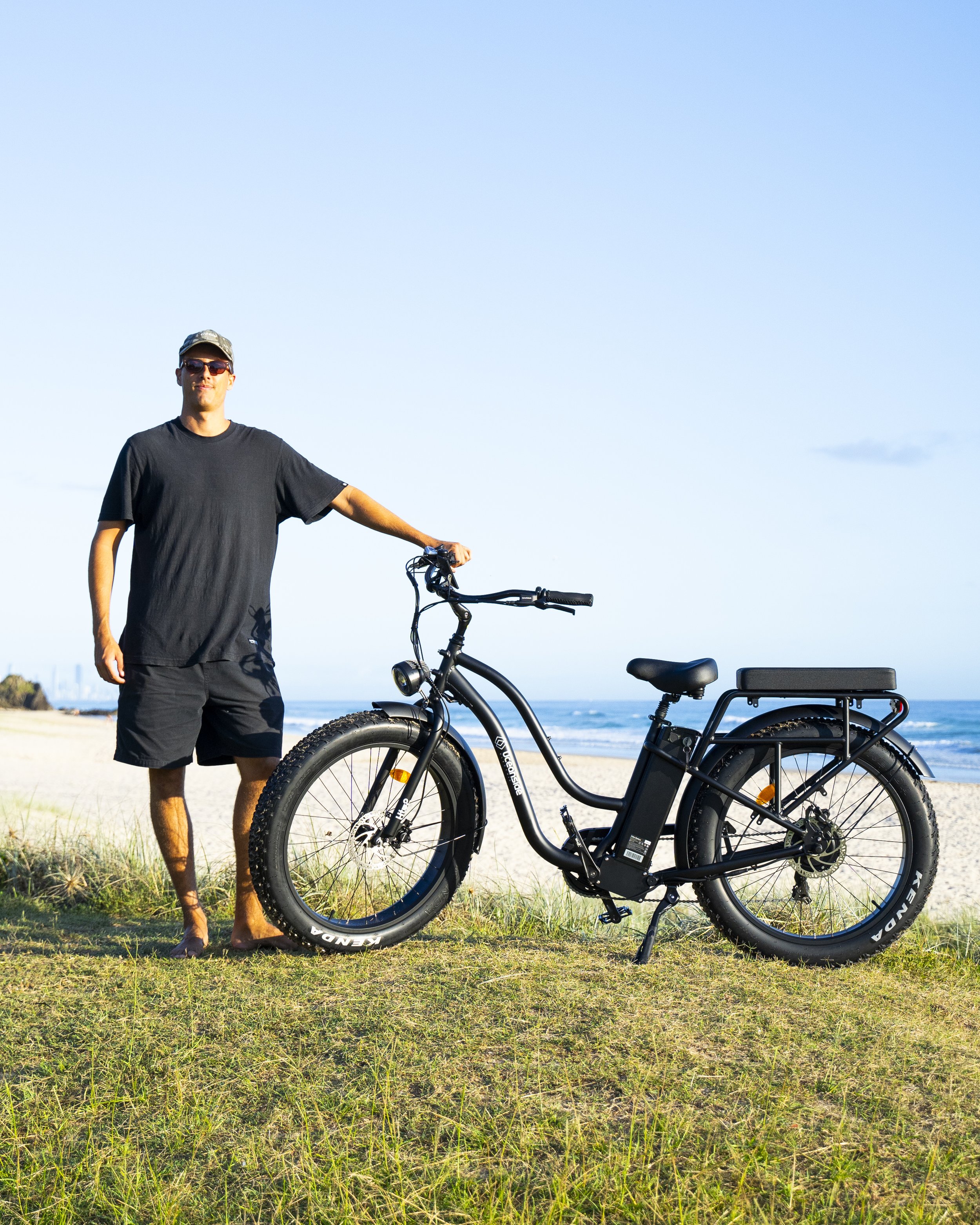 Man standing on grass next to an electric bike on a beach with ocean in the background.