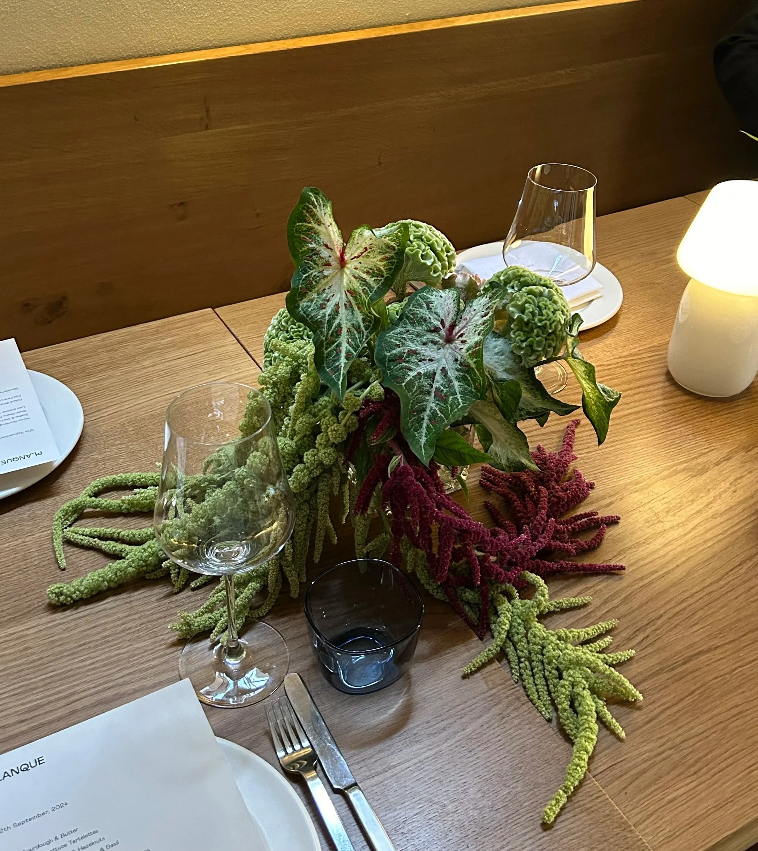 Table setting with a centerpiece of green and burgundy amaranthus on a wooden table, including glasses, white plates, cutlery, and a white lamp.