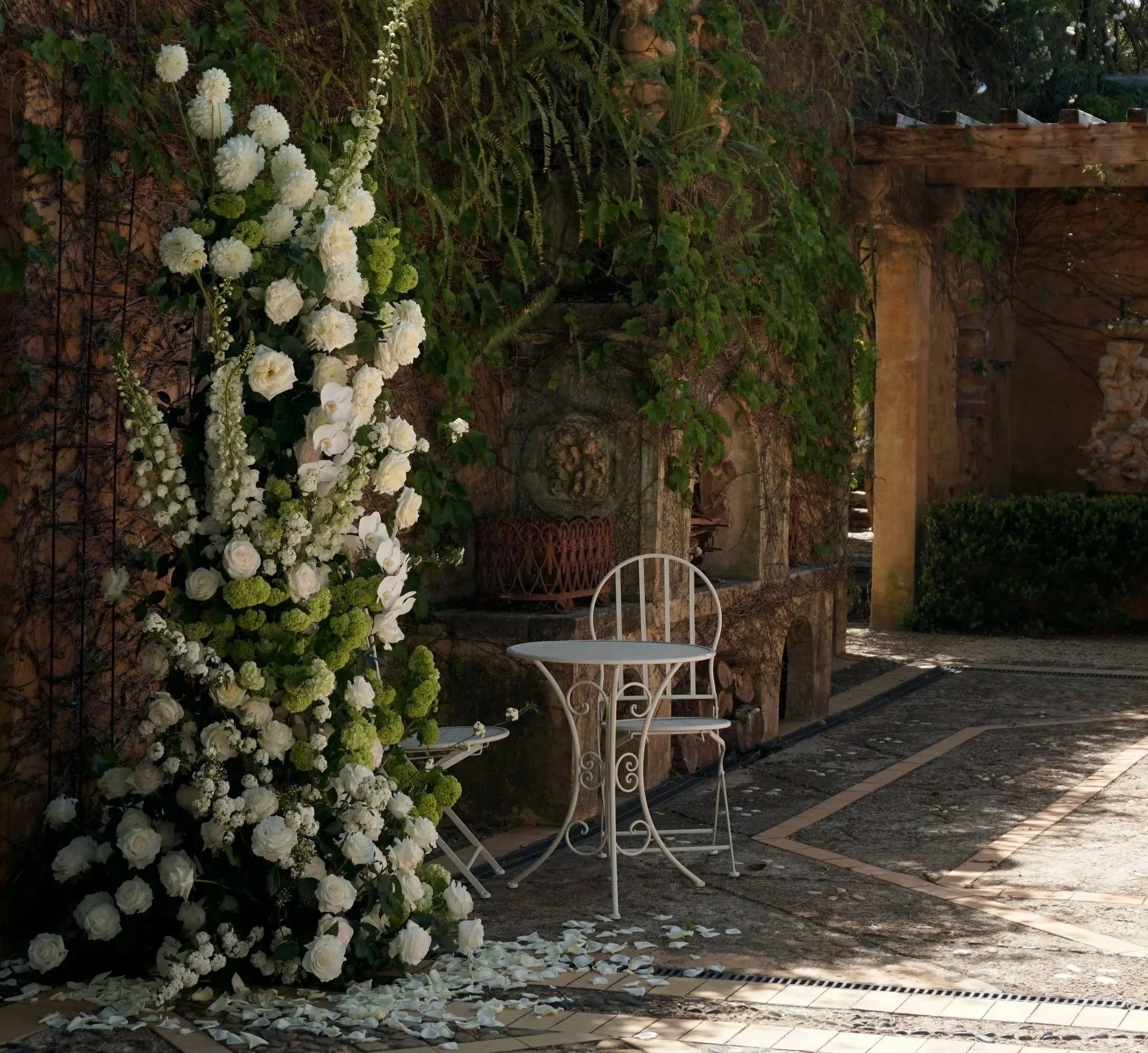 A white metal table with a matching chair, adjacent to a large floral arrangement of white roses, served with rose petal confetti on the ground, set in an outdoor courtyard with stone and brick features, greenery, and sculptures in the background.