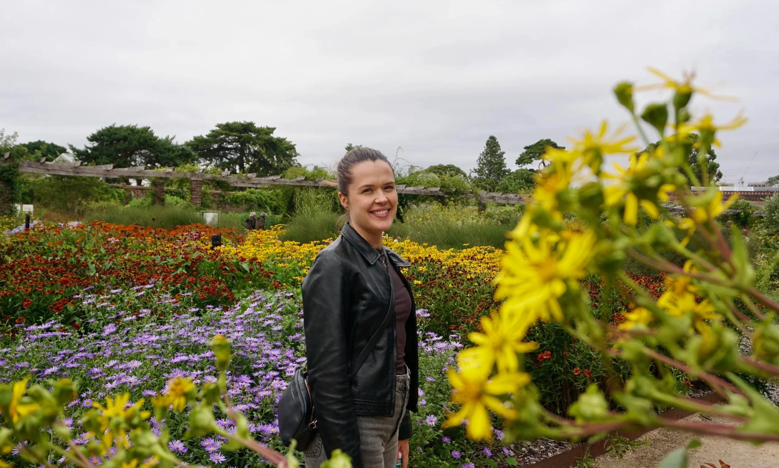 Grace smiling at the camera in a garden with colorful flowers and trees under an overcast sky In London