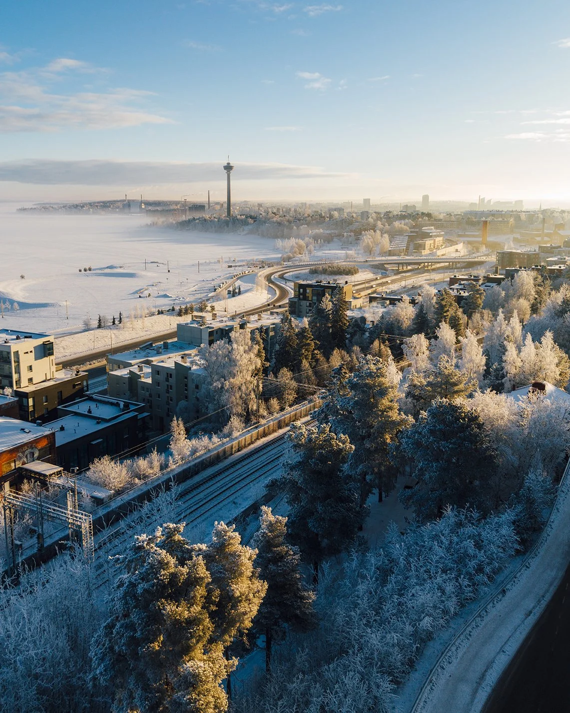 A snow-covered cityscape with trees, buildings, and a highway, with a foggy background and a tall tower in the distance.