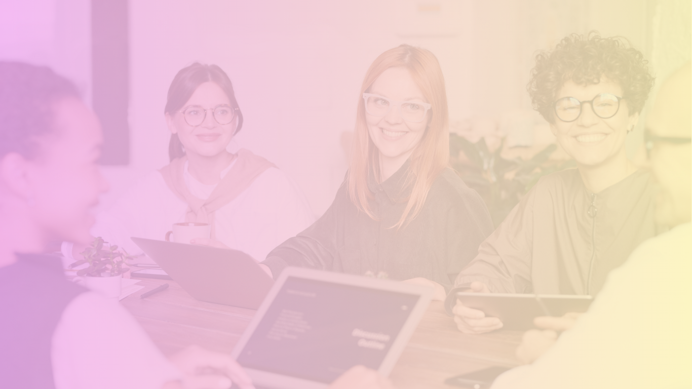 Group of five women sitting at a table with tablets, smiling and having a discussion in an office.