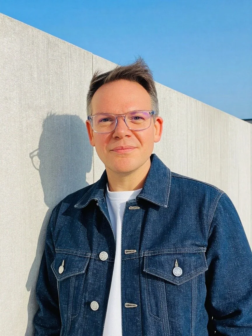 Adam Goodman-Smith. A man with glasses and light brown hair sitting at a wooden table, smiling, with a window and wooden wall behind him.