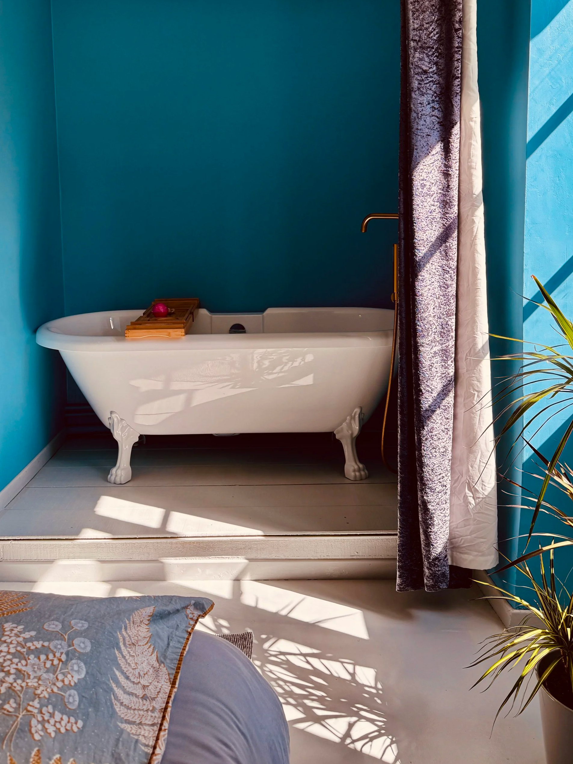 A white clawfoot bathtub with a wooden bath tray holding a pink bath bomb, against a blue wall with a dark textured curtain on the right side, with sunlight casting shadows on the floor and a plant in the lower right corner.