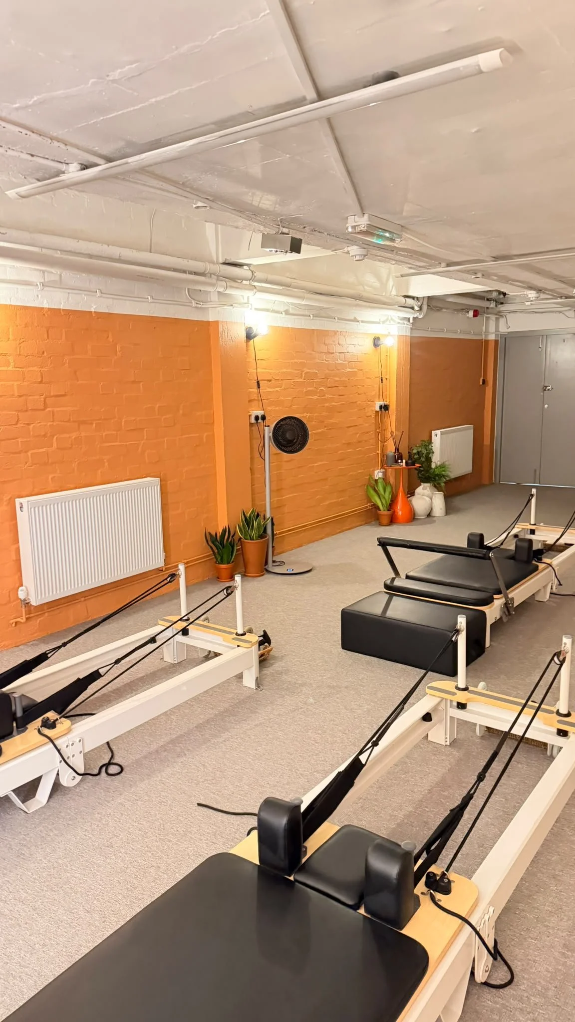 Exercise studio with reformer pilates machines, green plants, orange and white vases, a fan, and a brick wall painted orange. The ceiling has white pipes and fixtures, with wall-mounted lights.
