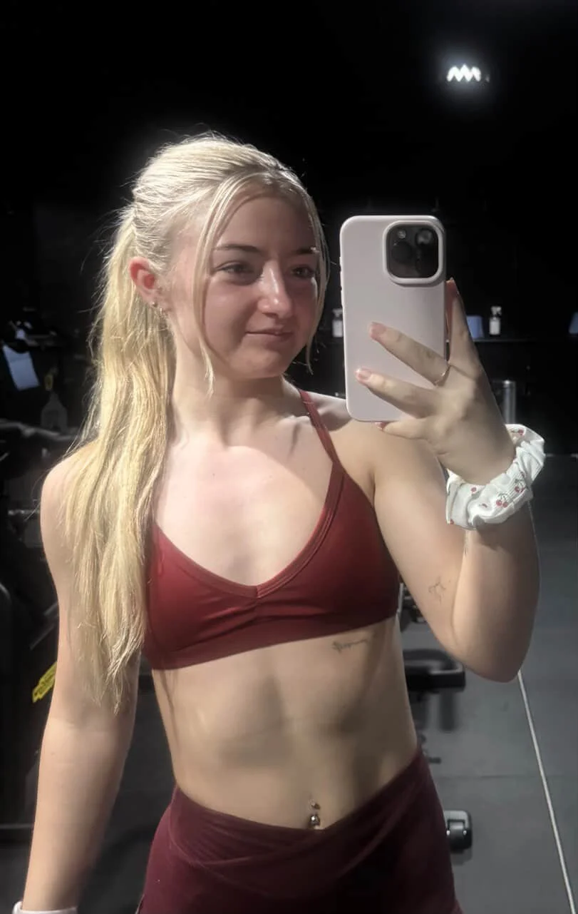 Young woman taking a mirror selfie in a gym, wearing a red sports bra and matching shorts, with a scrunchie on her wrist and a navel piercing.