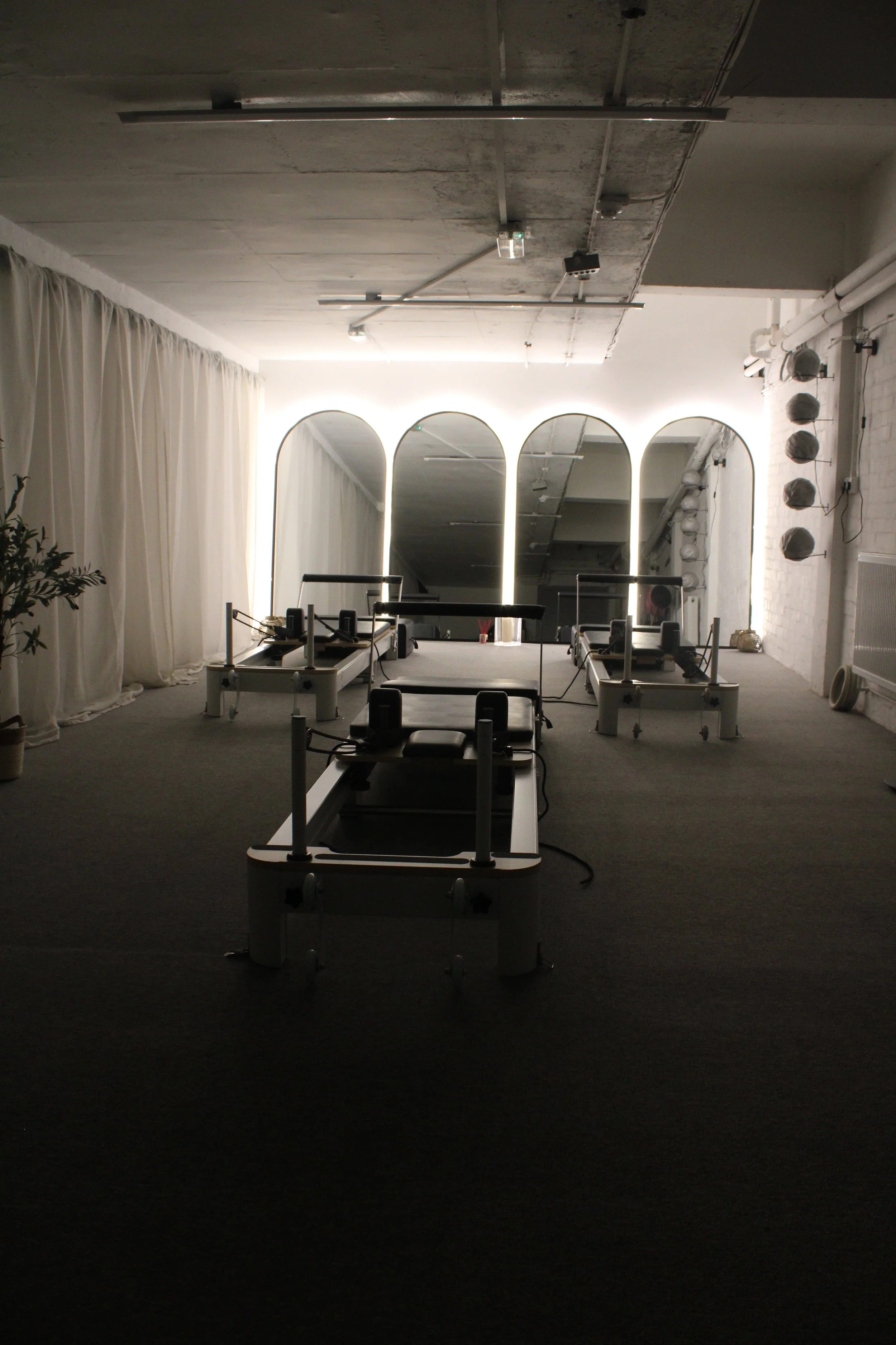 A Pilates studio with reformer machines arranged in a row facing large mirror panels with backlit arches, white curtains on the left, and white brick walls with a row of reformers on the right.