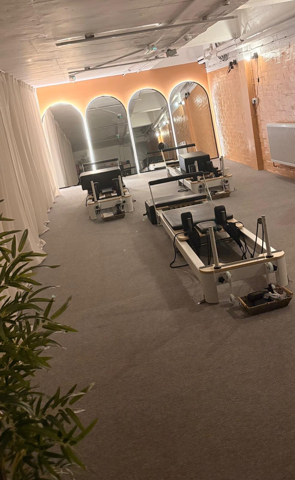 The image shows a spacious room with three Pilates reformer machines arranged in a row on a grey carpeted floor. The room has a warm orange accent wall with three large, arched mirrors and backlit lighting. To the left, there are white curtains, and on the right, an exposed brick wall with white pipes and lighting fixtures. Some greenery is visible in the lower left corner.