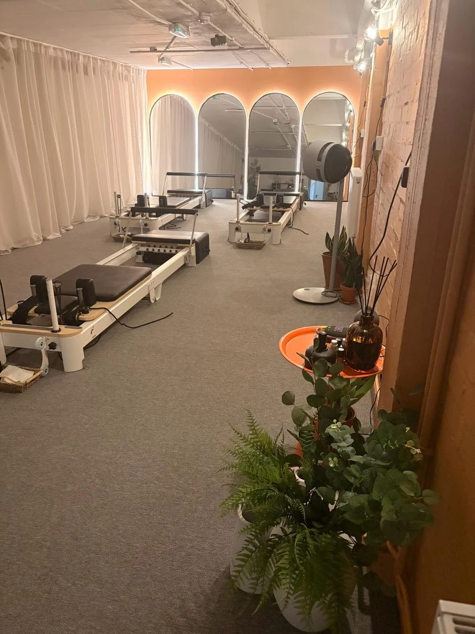 A wellness or Pilates studio with reformer machines, potted plants, a fan, and a mirror wall with arch-shaped frames, illuminated by soft lighting.