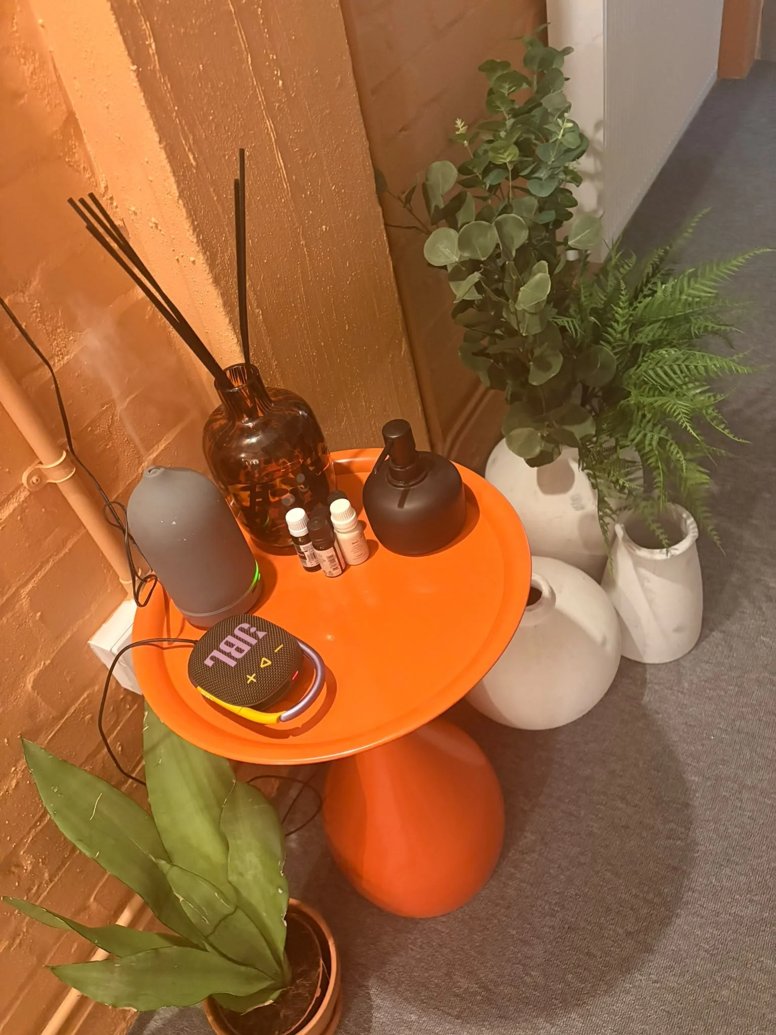 An orange table with a large green leafy plant in a white pot, a smaller leafy plant in a white pot, a brown glass bottle, a black diffuser, several small bottles, and a black and yellow portable speaker. The table is against a brown brick wall, and the scene is lit warm.