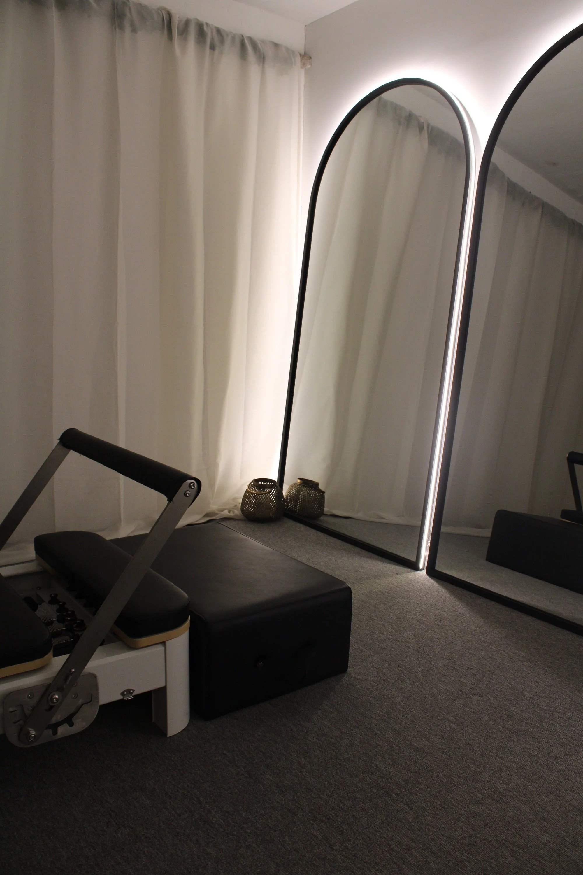 A modern room corner with white curtains, a large upright mirror with black edges, two decorative candle holders, a black ottoman, and a piece of exercise equipment.